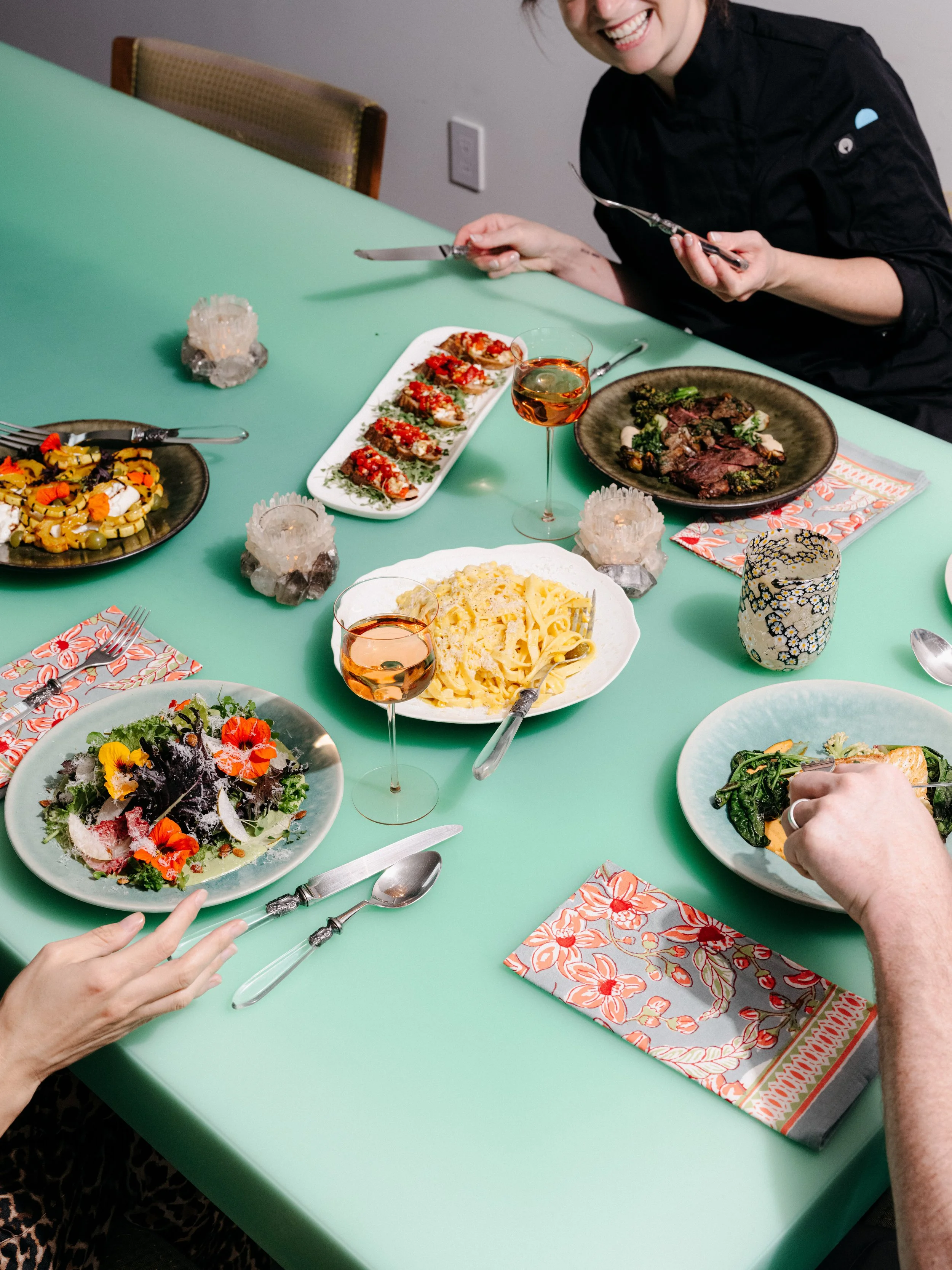 People enjoying a meal at a table with various dishes including pasta, salad, and meat, along with wine and decorative candles.