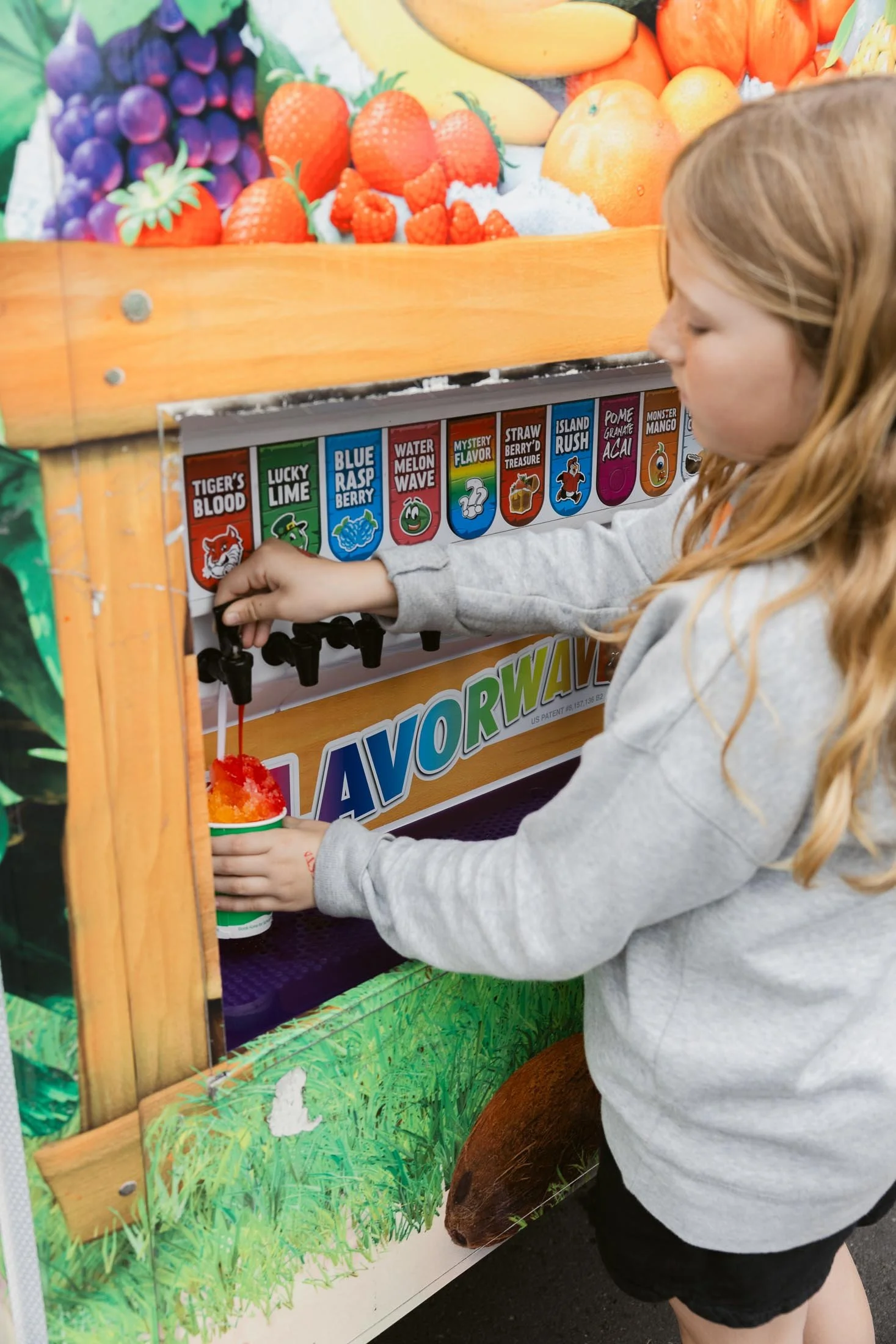 A young girl with red hair in a gray hoodie and black shorts is filling a cup with red and orange snow cone from a colorful snow cone stand decorated with images of fruits and animals.