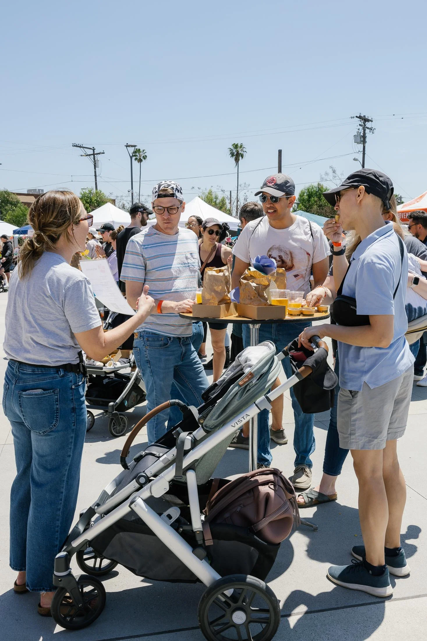 Group of people at an outdoor event under a clear blue sky, standing around a table with food and drinks, some pushing strollers, with white tents and palm trees in the background.
