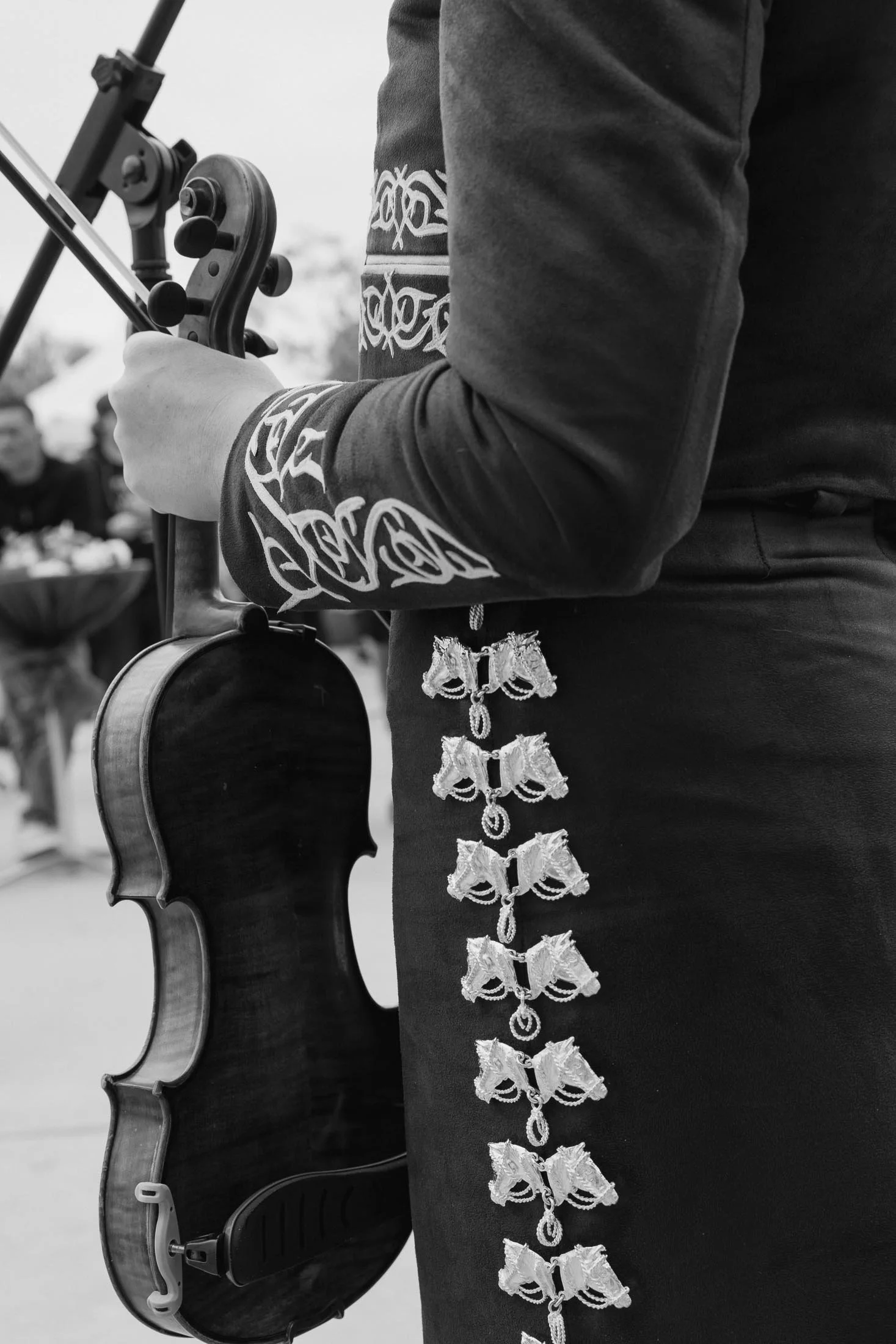 A person holding a violin, with a decorative jacket sleeve featuring embroidered detailing. They are at an outdoor event, with blurred people and food stands in the background, and are adjusting the violin.
