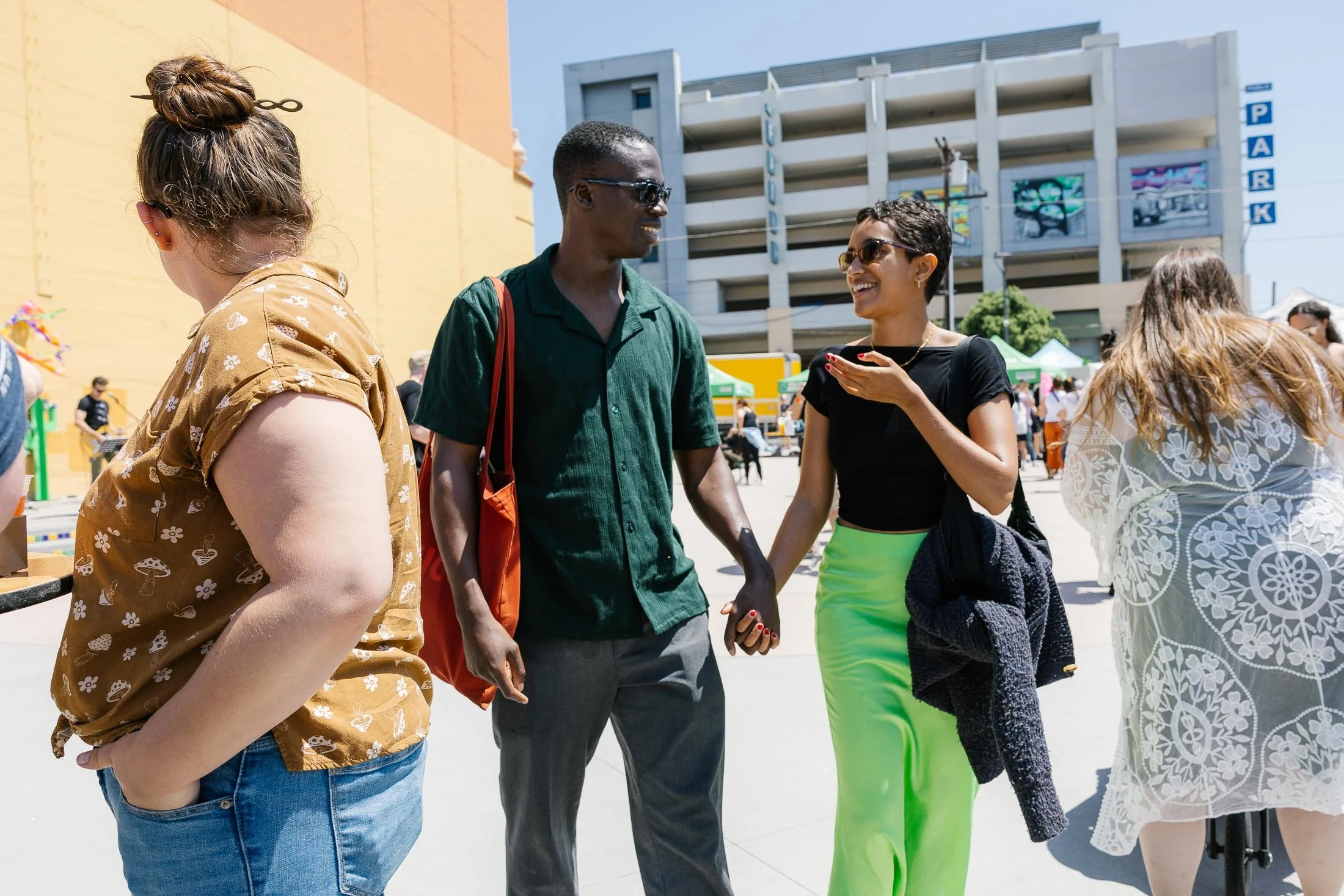 Two women and a man holding hands and smiling in an outdoor urban setting during daytime, with a building, tents, and people in the background.