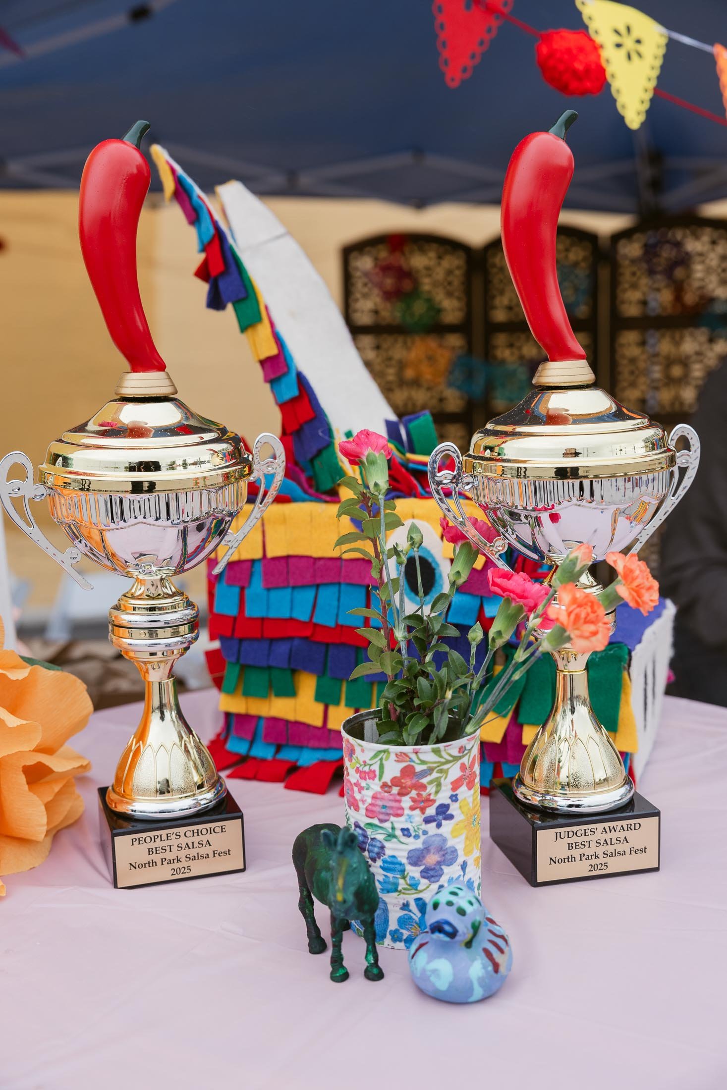 Two silver trophies with red chili pepper handles, colorful papel picado banners in the background, and a vase with pink flowers on a pink tablecloth. There are small decorative figurines of an elephant and a bird in front of the vase. The trophies a