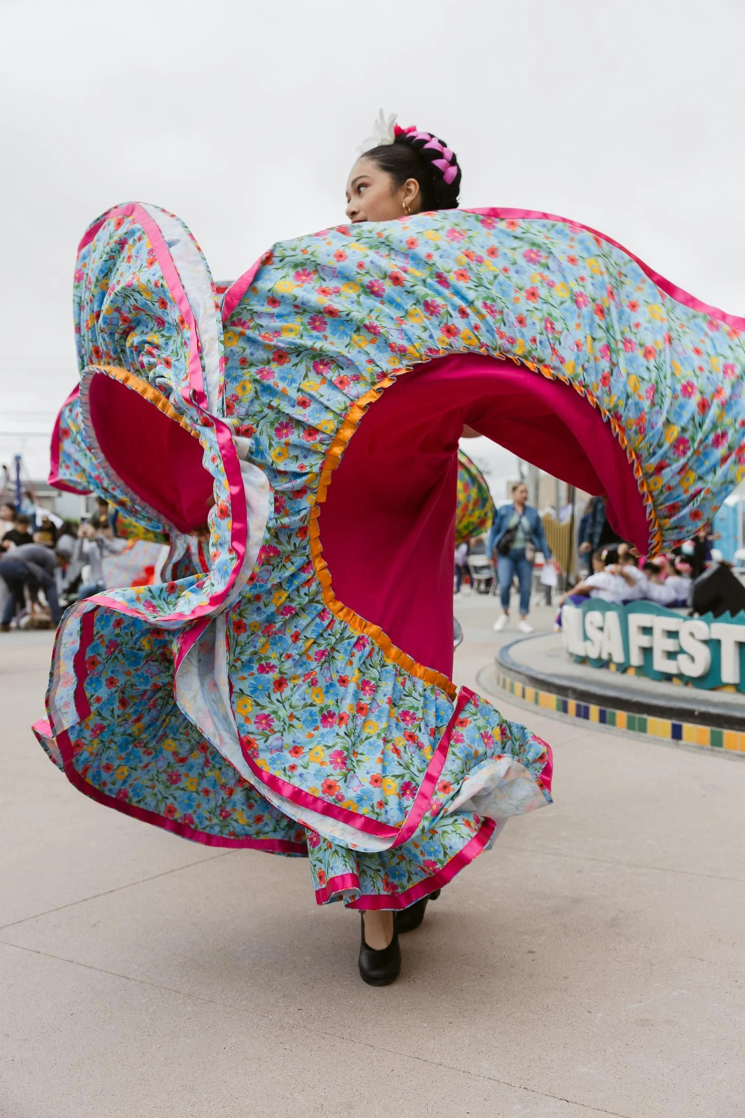 A woman dancing in a colorful, floral dress at an outdoor festival, with blurred crowd and a sign reading 'LSA FEST' in the background.