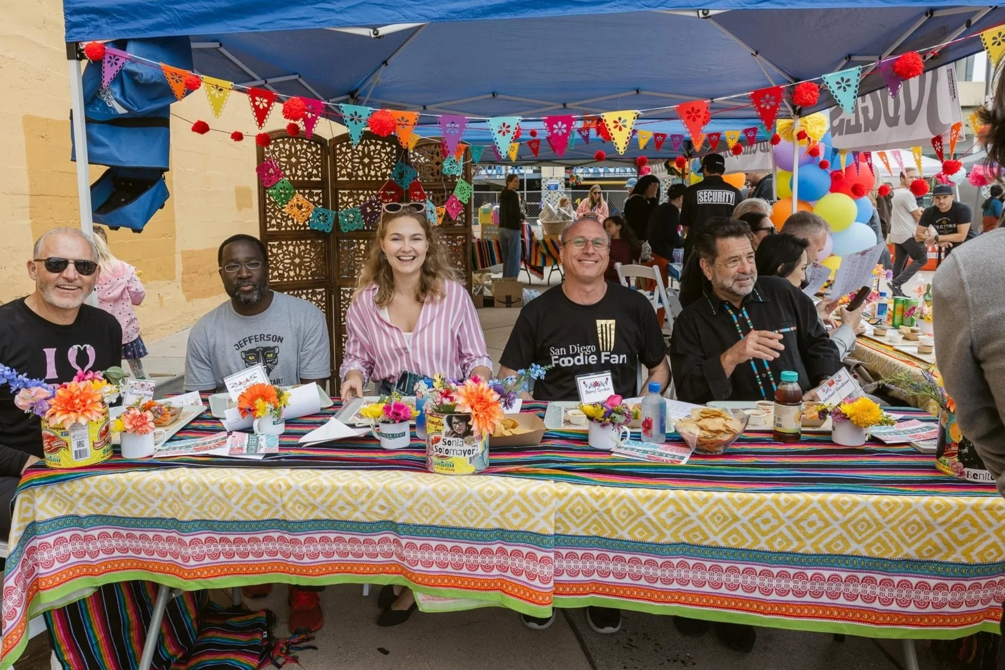 People sitting at a decorated table outdoors during a festive celebration, with colorful banners, balloons, flowers, and food on the table.