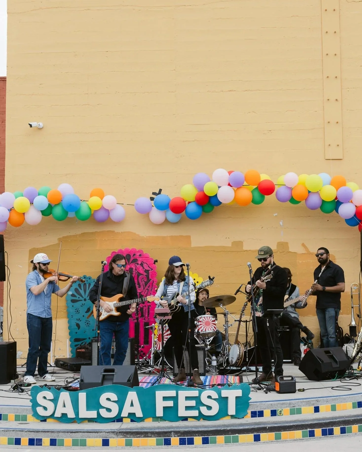 A live band performs on an outdoor stage with a colorful "Salsa Fest" sign, decorated with balloons and vibrant paper cutouts, in front of a yellow wall.
