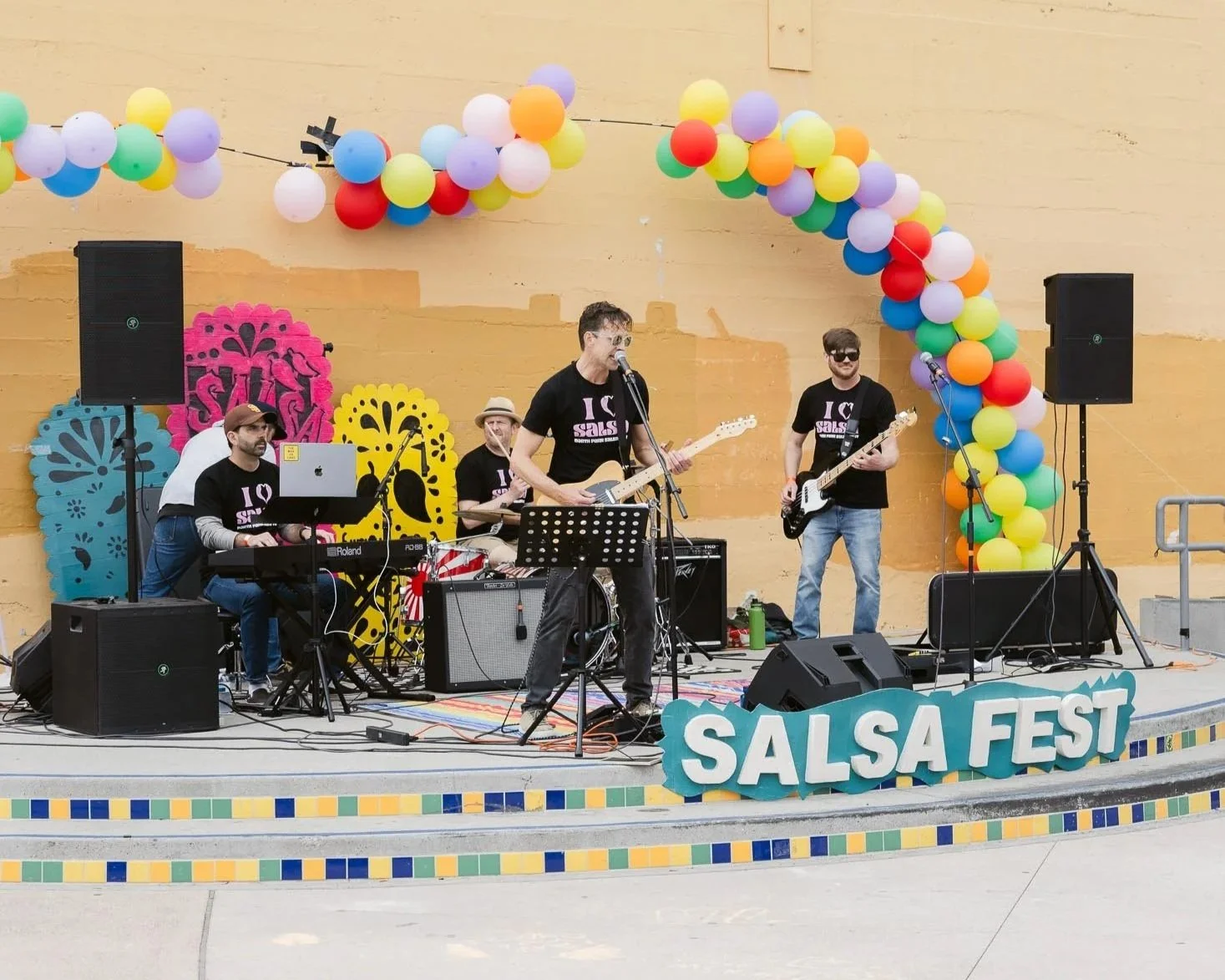 A band performing live at Salsa Fest with colorful balloon decorations and a large sign that reads 'SALSA FEST' in front of an orange and yellow wall.