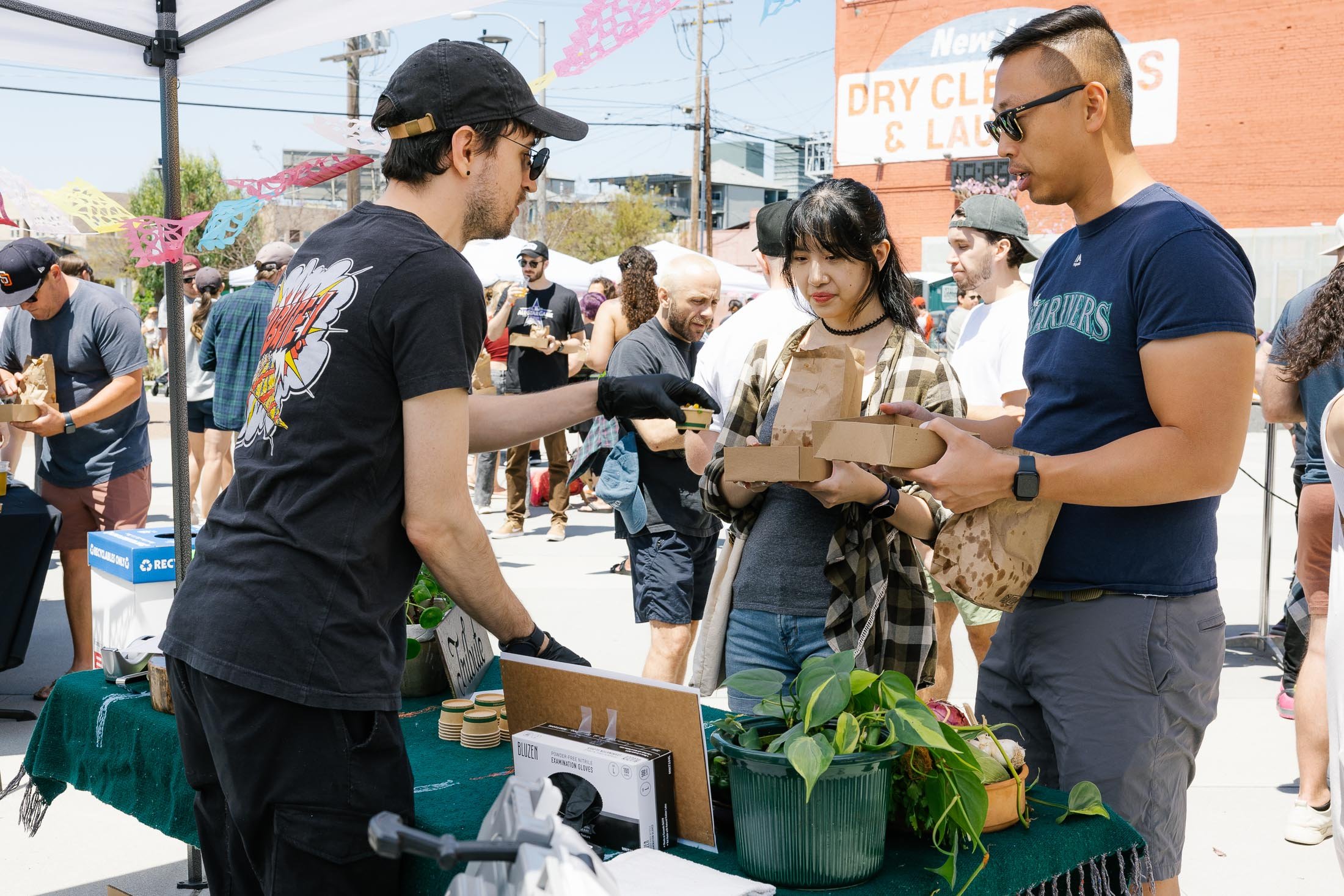 People at an outdoor market exchanging food, with a man handing a box to a woman, surrounded by other shoppers and market stalls.