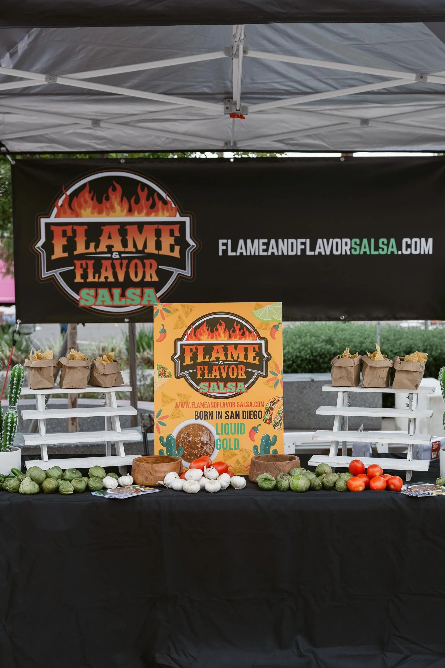 A booth with a black banner that says 'Flame & Flavor Salsa' and a website address, with a colorful sign promoting salsa made in San Diego and featuring vegetables like tomatoes, garlic, onions, and cucumbers displayed on the table along with paper b