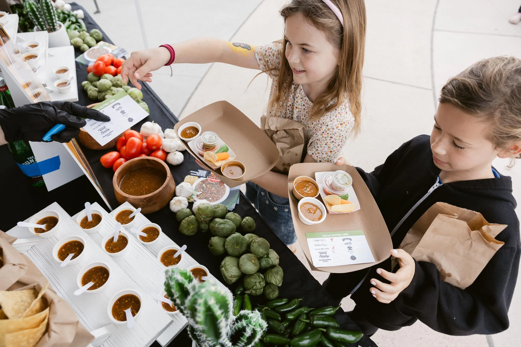 Two children at a market stand are receiving small cups of sauce or salsa from a vendor. The stand has fresh vegetables like tomatoes, jalapeños, and green vegetables, with some small containers of sauce and a tasting card.