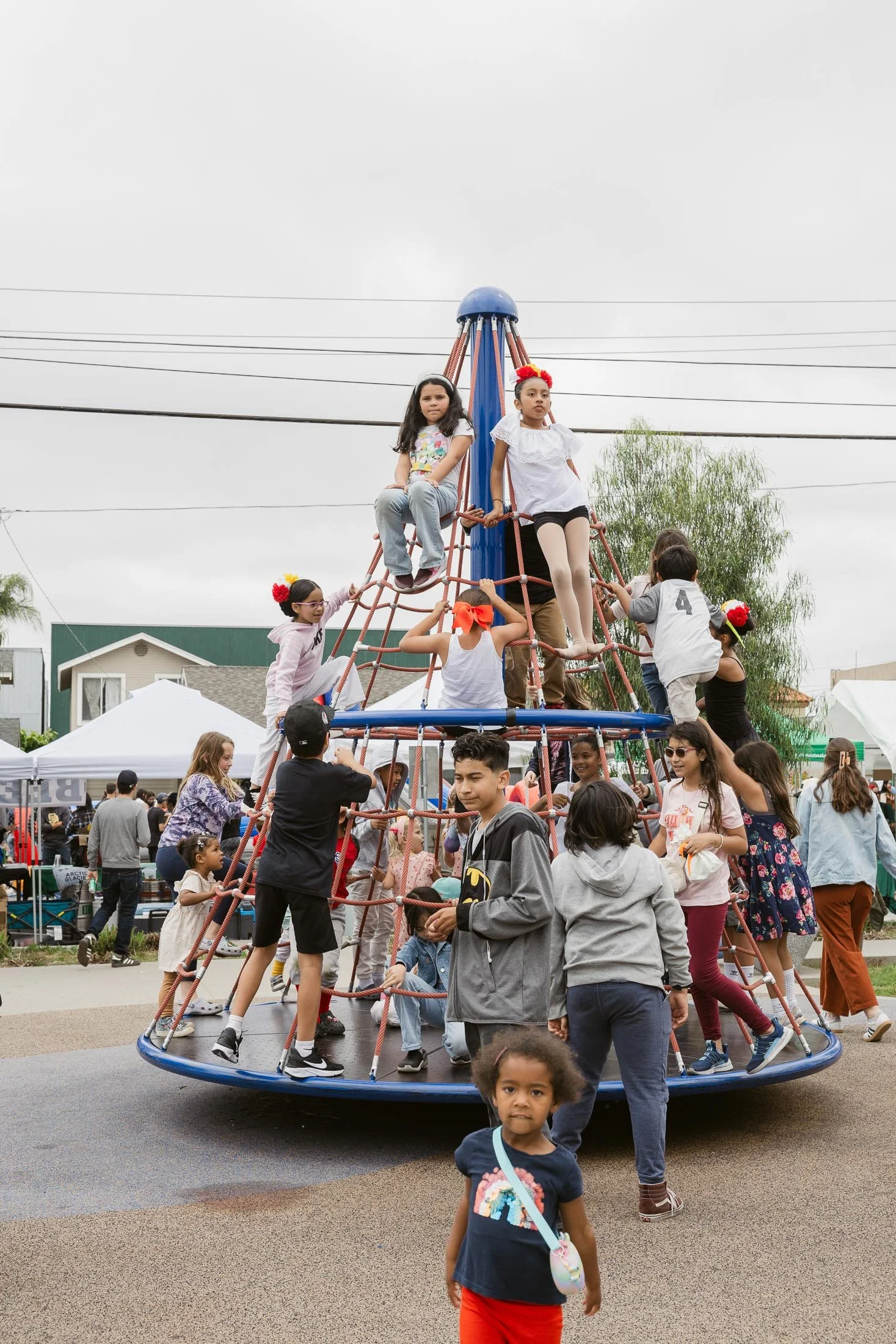 Children playing on a large, conical climbing structure at an outdoor event, with tents and people in the background on a cloudy day.
