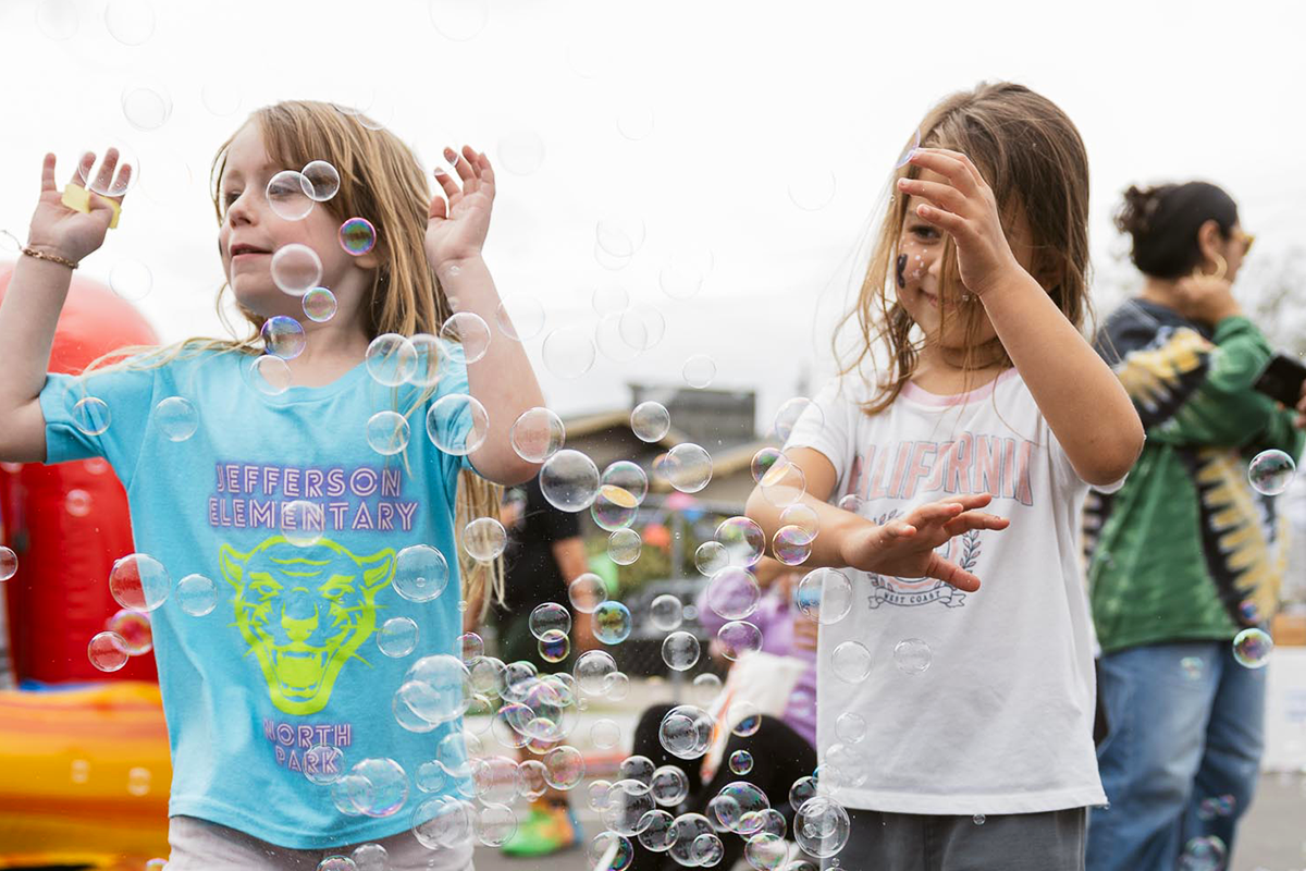 Two young girls playing with soap bubbles outdoors on a cloudy day, with other children and a person in the background.