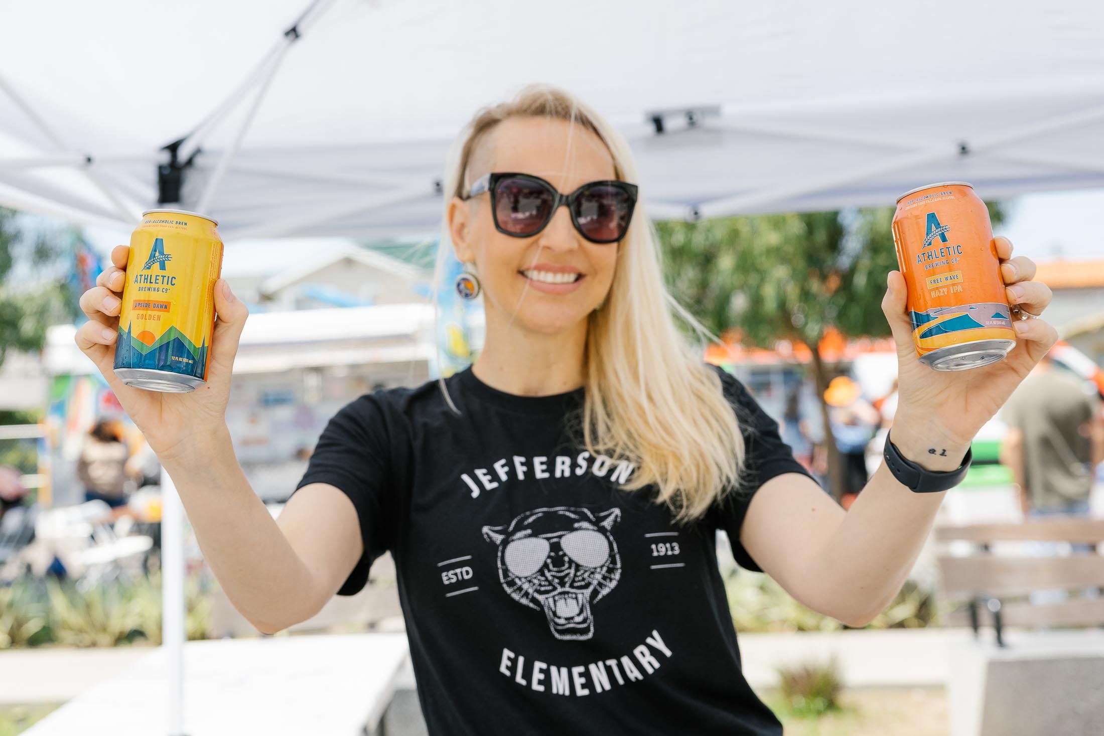 A woman with blonde hair wearing sunglasses and a Jefferson Elementary T-shirt, smiling and holding two cans of Athletic Brewing Co. non-alcoholic beer.