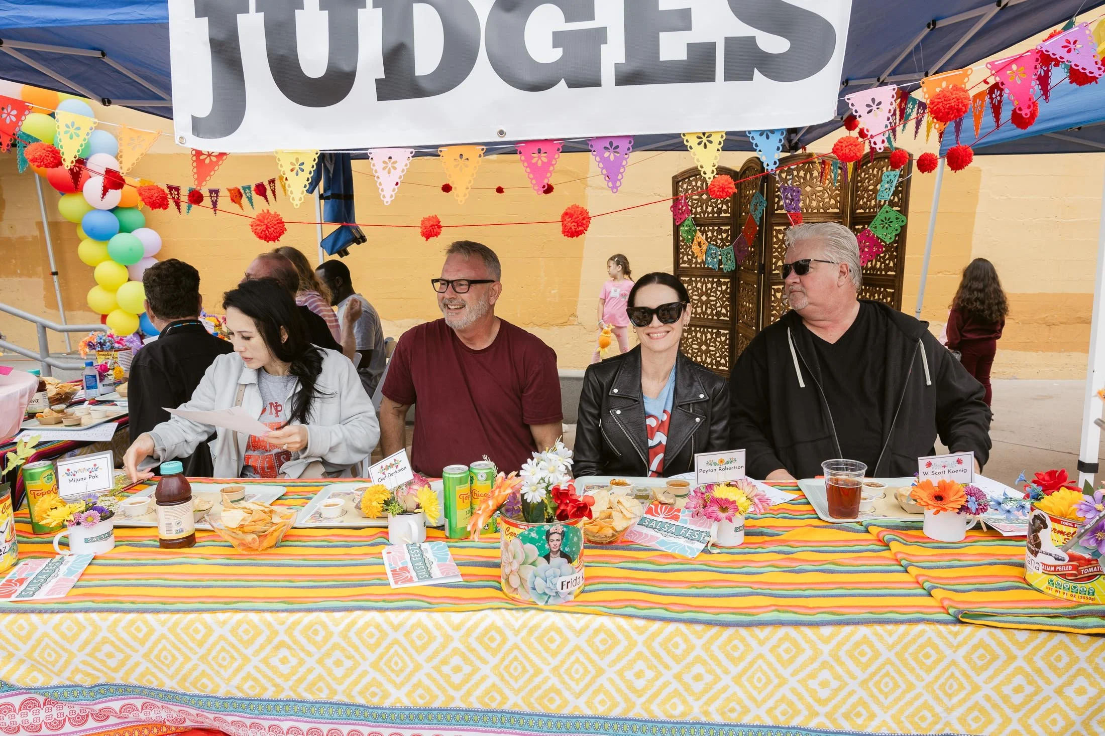 People sitting at a long table decorated with colorful tablecloth, flowers, and party items under a canopy labeled 'JUDGES'. There are four people sitting in front, smiling and wearing casual clothes. Behind them, there are more people and children, 