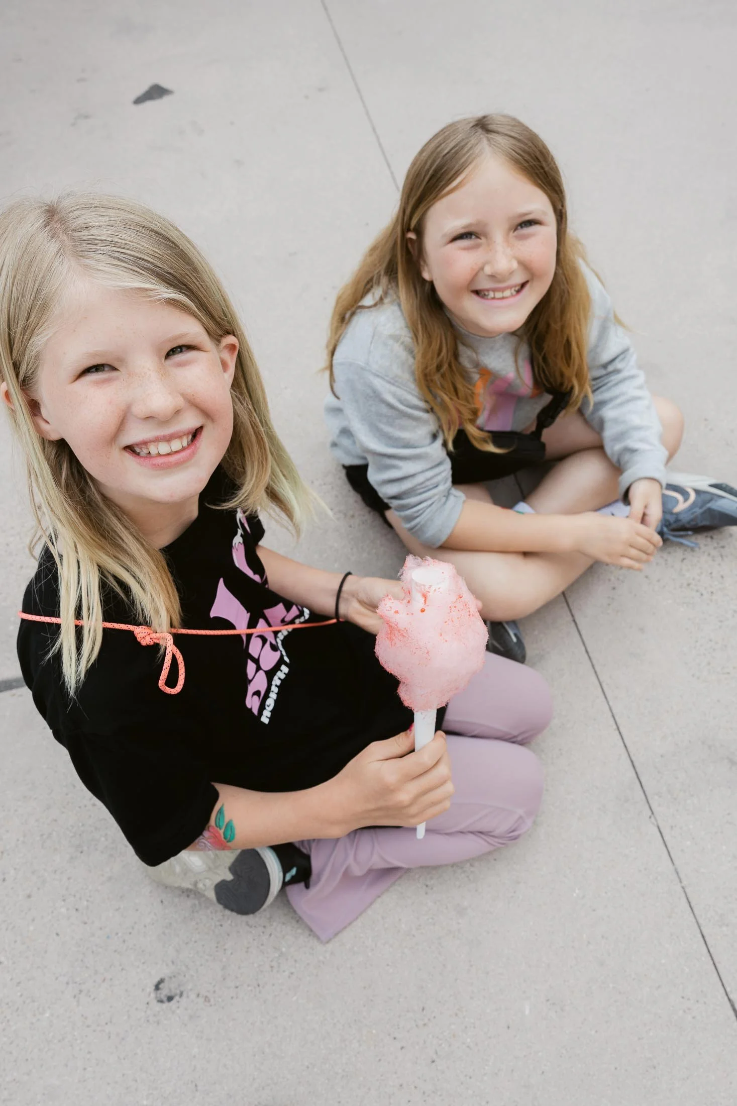 Two young girls sitting on the ground smiling, one holding pink cotton candy. They are outdoors on a concrete surface.