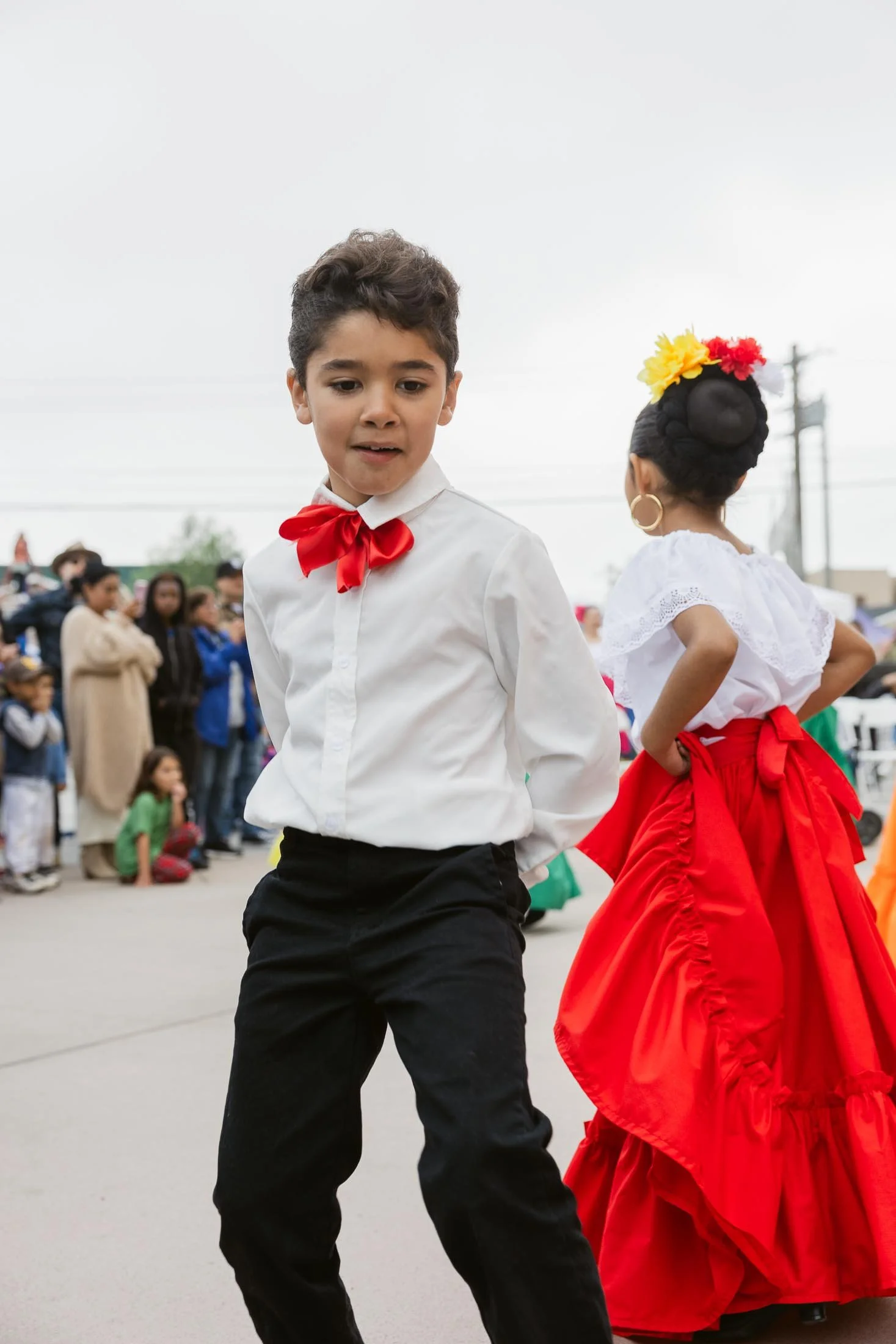 A young boy wearing a white shirt with a red bow tie dances at a cultural event. In the background, a girl dressed in a traditional red and white dress with a floral headpiece also participates, while a crowd of people watch.