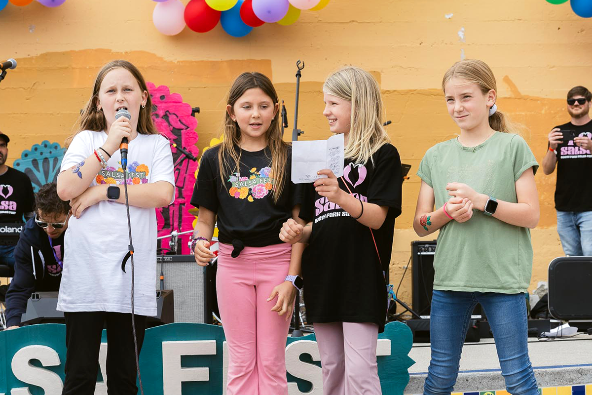 Four young girls standing on a stage during a salsa festival, with colorful balloons overhead. One girl is speaking into a microphone, while others are listening and smiling. There are some people and musical equipment in the background.
