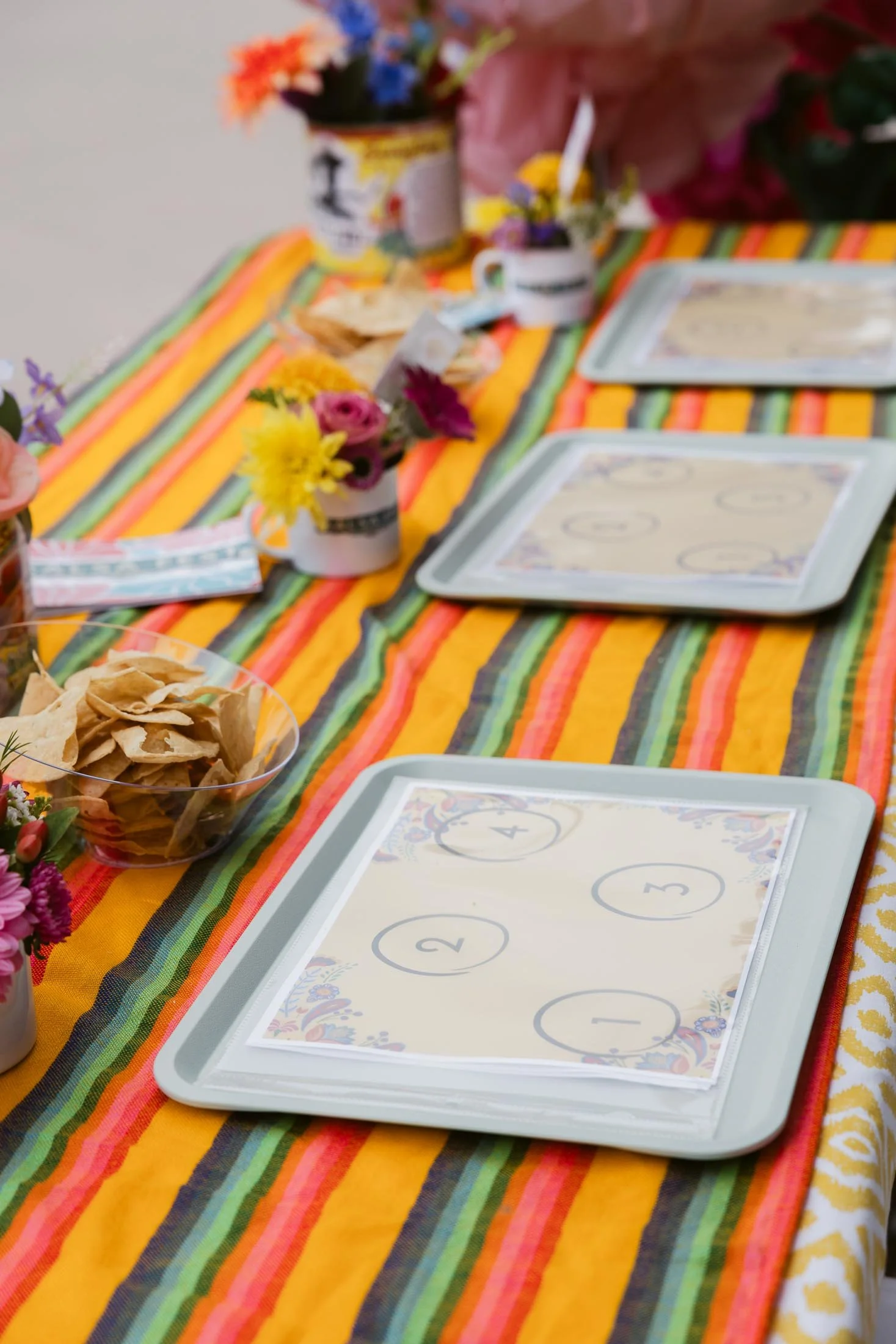 A table decorated with a colorful striped tablecloth featuring orange, yellow, green, and black stripes, set for a meal with four trays numbered 1 through 4, small vases with colorful flowers, plates, and bowls of chips and snacks.