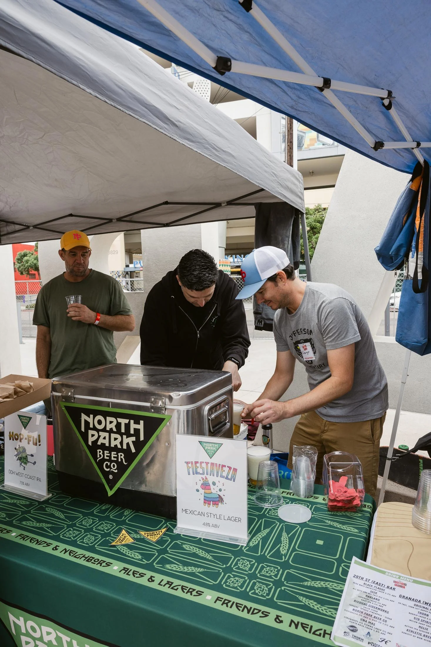 Three men working at a beer stand under tents at an outdoor event. One man is pouring drinks, while the other two observe. The stand is labeled North Park Beer Co and offers Mexican style lager called Fiesta Weza. There are plastic cups and taps on t