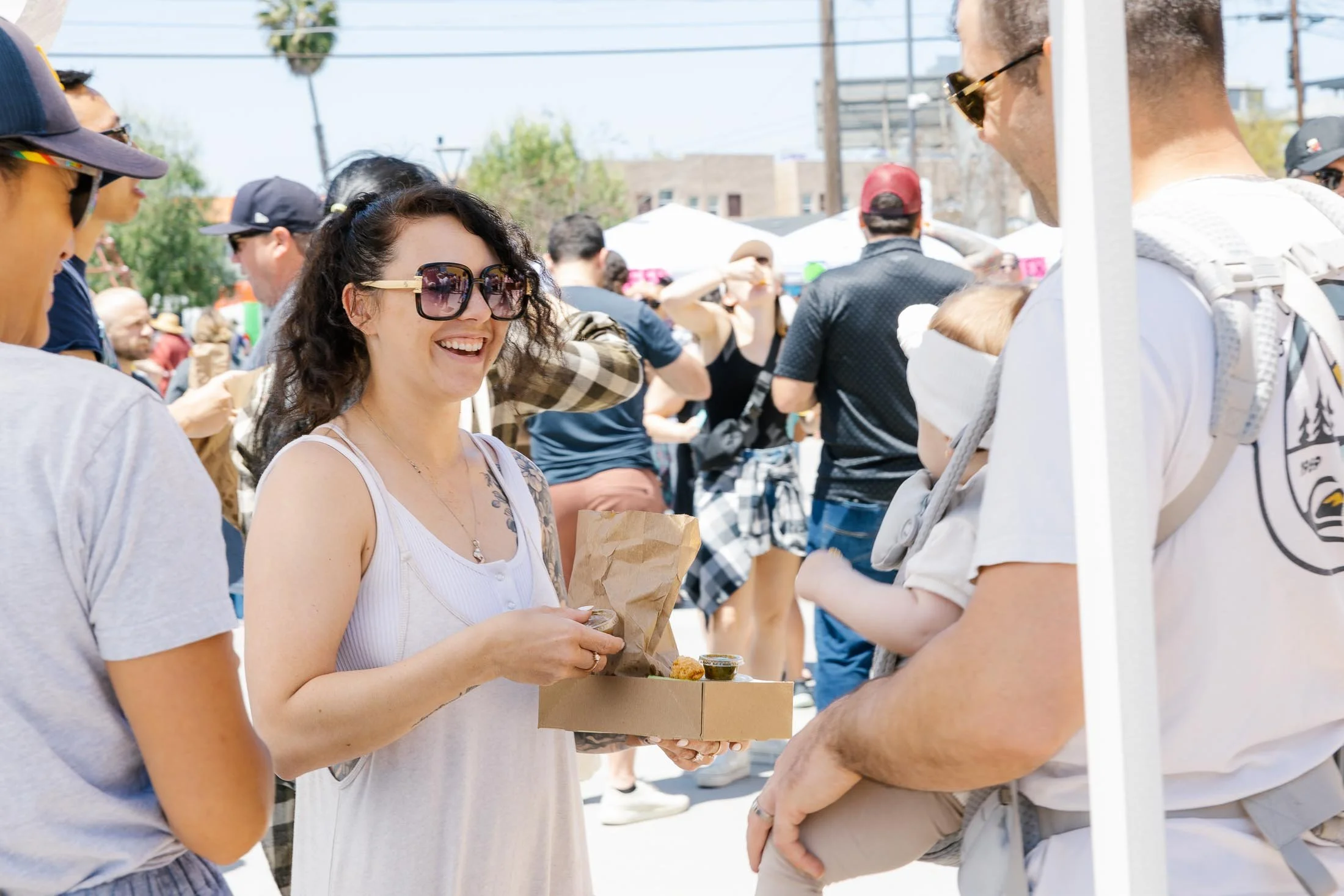 A woman with curly hair and sunglasses smiling while handing food to a man with a child in a baby carrier at an outdoor event.