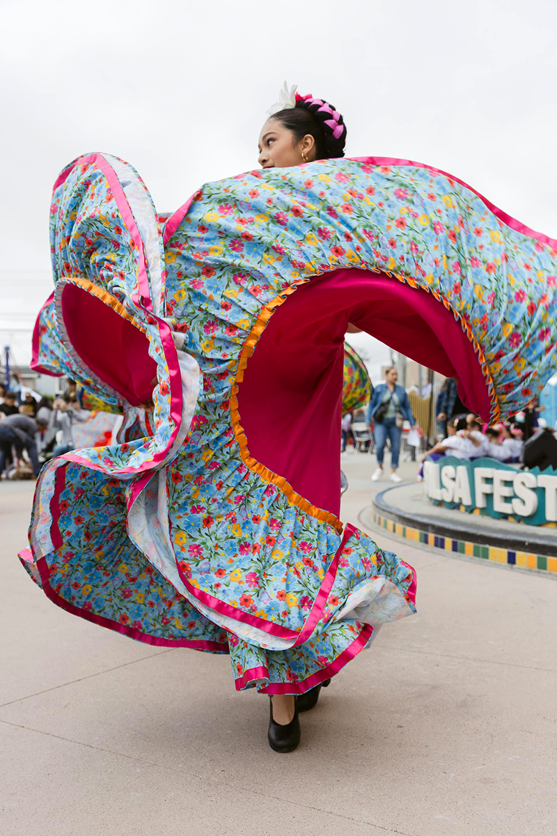 A woman in traditional Mexican attire performing a folkloric dance, with a colorful floral dress and a white flower in her hair, at an outdoor festival.