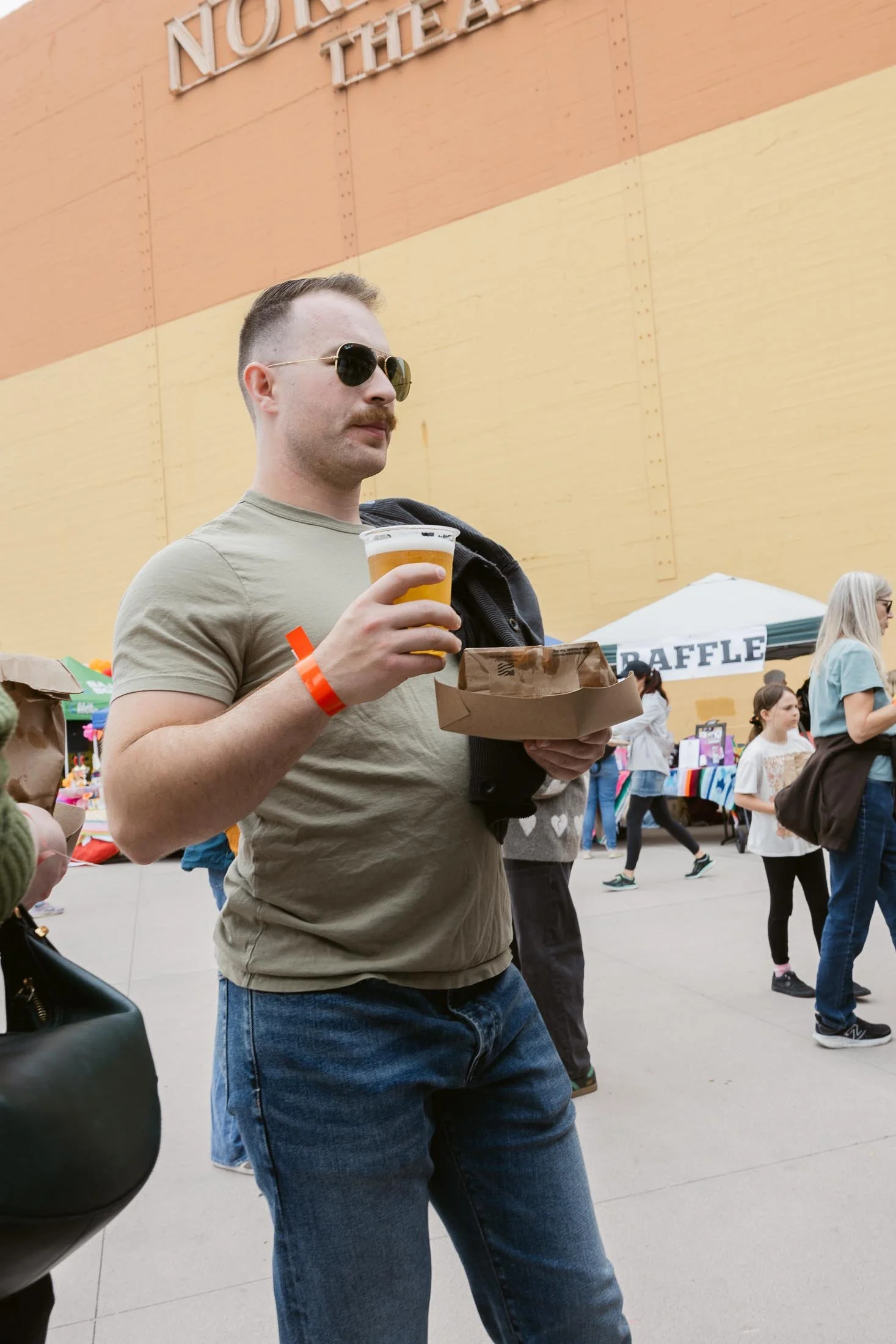 A man with a mustache and sunglasses holding a beer and a food tray at an outdoor market or festival.