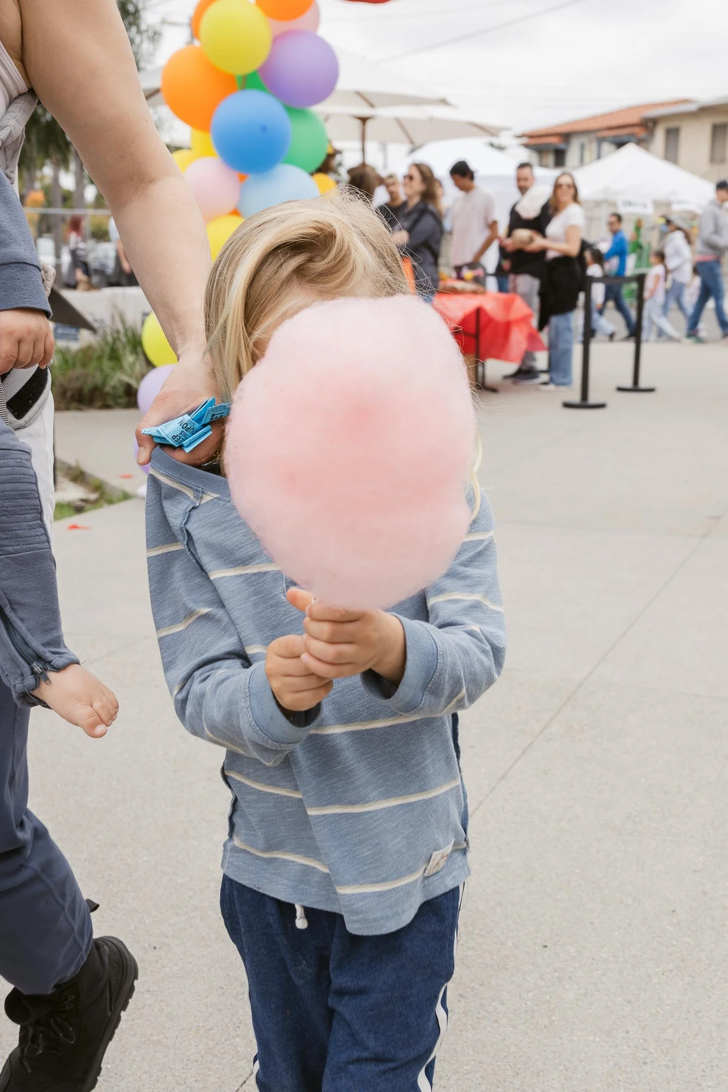 Child holding pink cotton candy in front of face at outdoor event with balloons and groups of people in background.