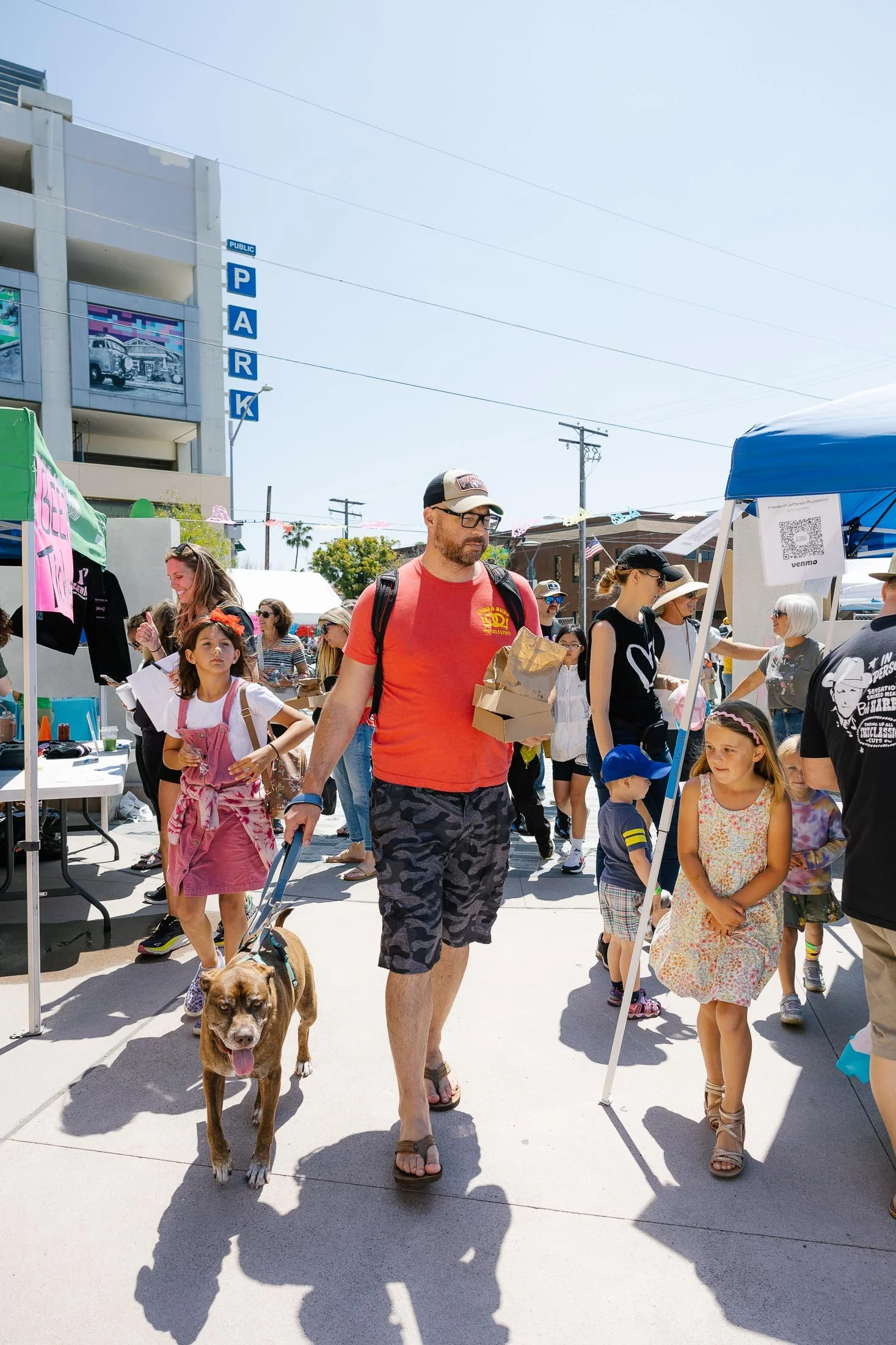 People walking at an outdoor market on a sunny day, with a man in a red shirt walking a dog, surrounded by children and vendors.