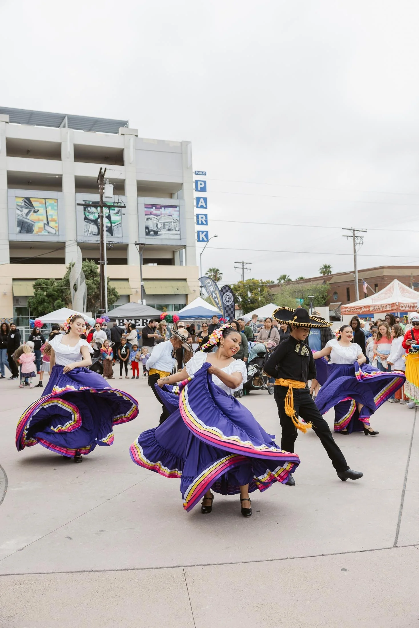 Mexican folkloric dancers performing outdoors during a festival, with a crowd watching and vendor tents in the background.