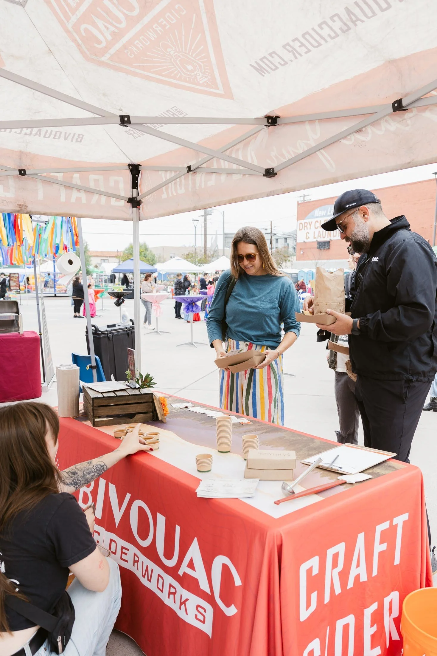 People at a craft and cider booth under a large tent at an outdoor event, with colorful decorations and other vendors in the background.