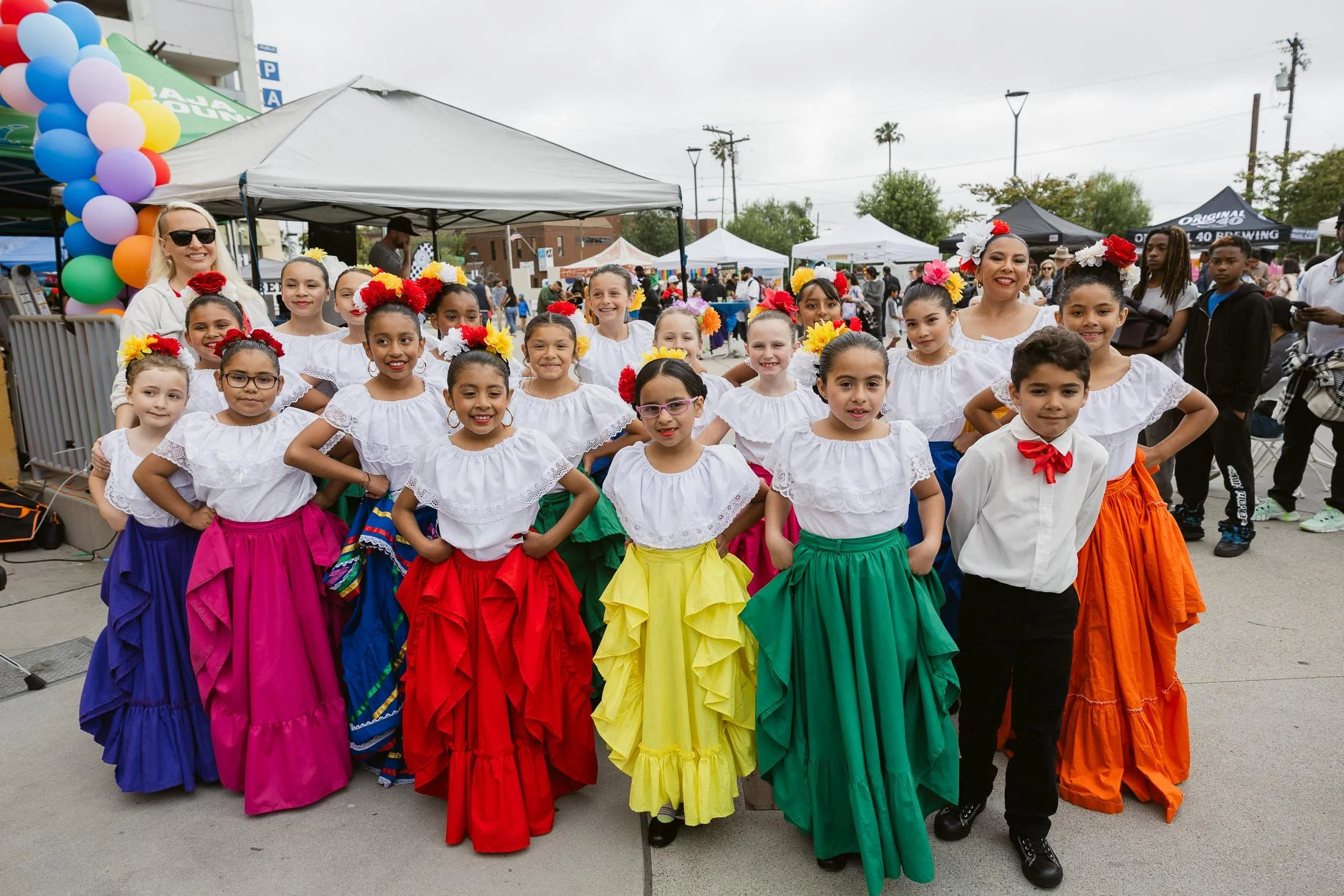 Group of young girls in traditional Mexican attire, colorful skirts and white blouses, standing outdoors at a cultural event or festival.