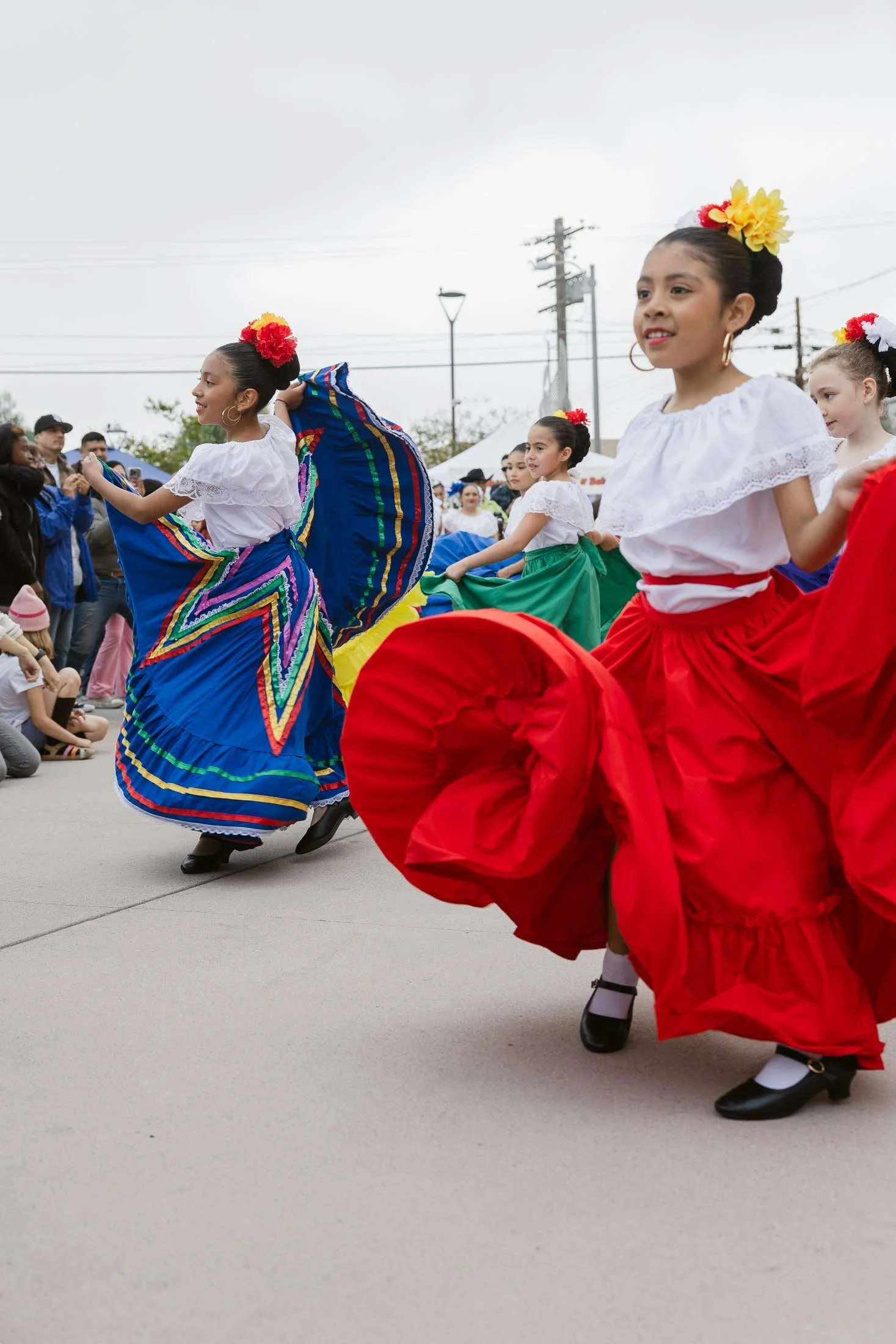 Young girls in traditional Mexican dresses dancing during a parade, with colorful skirts, flowers in hair, and spectators watching.