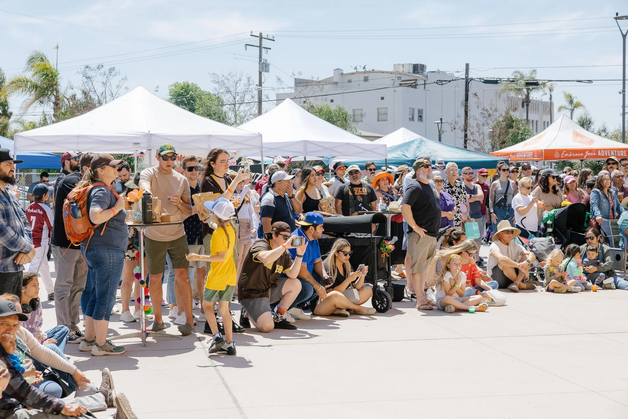 Crowd of people gathered at an outdoor event with white tents, some sitting on the ground, some standing, many taking photos or videos, on a clear, sunny day.