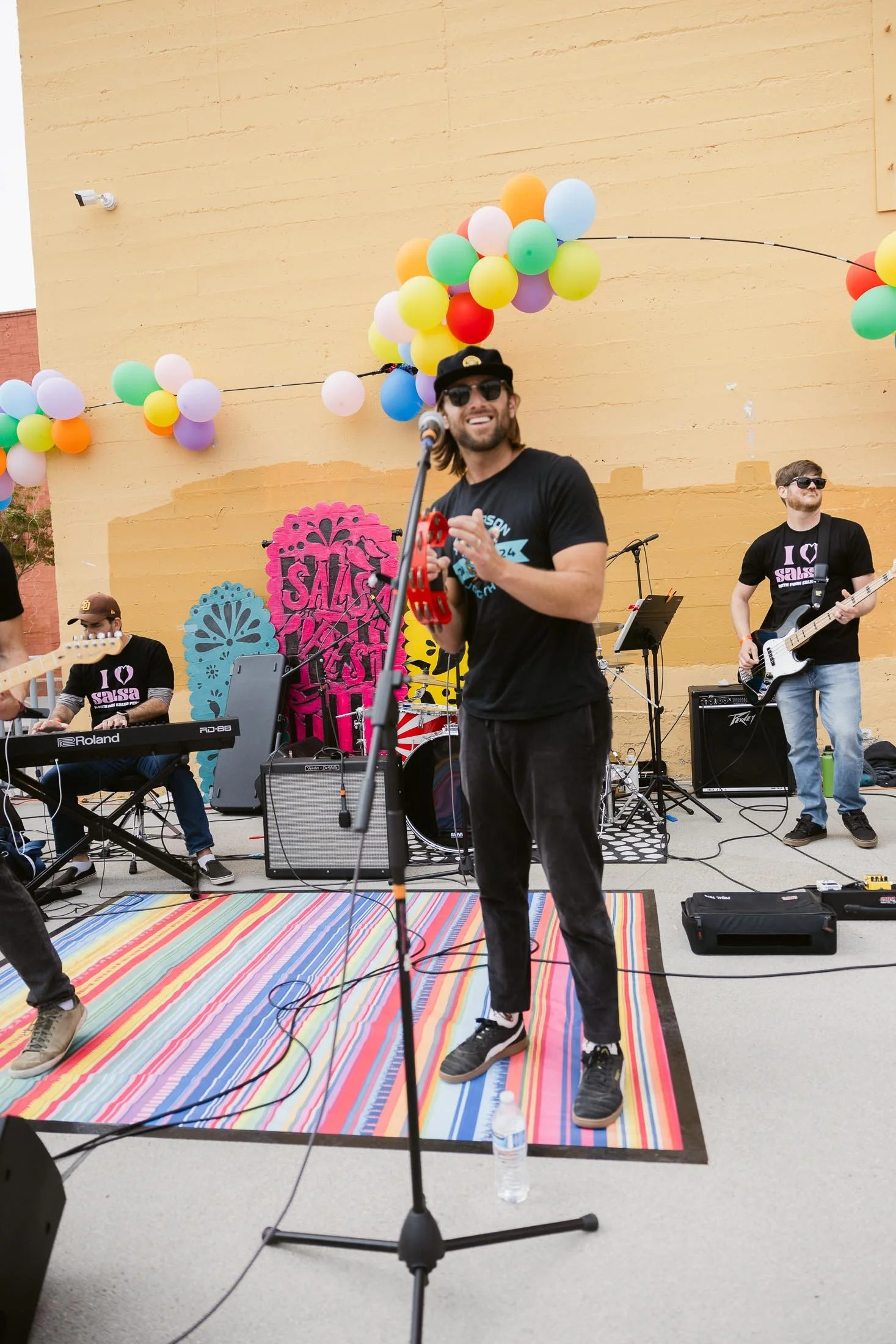 A band performs outdoors under colorful balloons and decorations, with a man in sunglasses holding a microphone and tambourine, smiling. Other band members are playing instruments against a yellow wall decorated with pink, blue, and yellow designs.