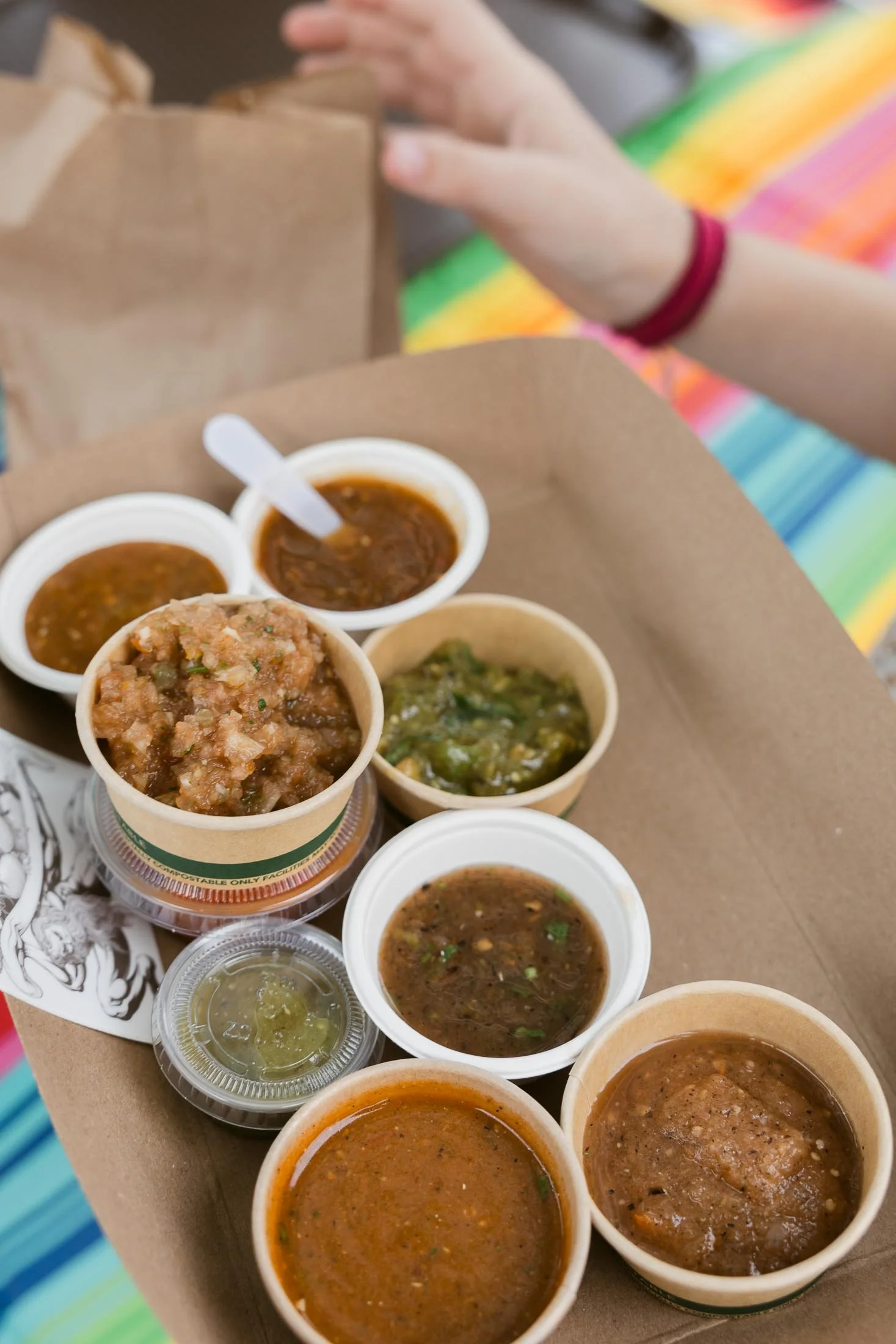Multiple small cups of assorted salsas and dips on a brown tray, with a person's hand and rainbow-colored fabric in the background.