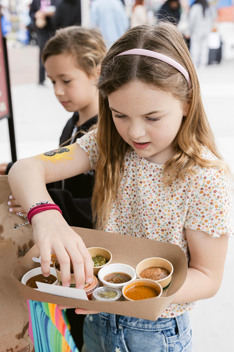 A young girl with a pink headband and floral shirt looking at a tray of small bowls with various sauces and condiments at an outdoor event.