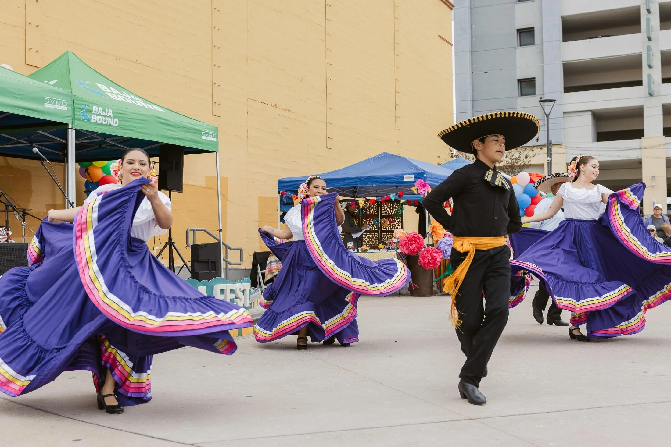 Mexican dancers performing in traditional attire at a cultural celebration with colorful decorations and live music setup.