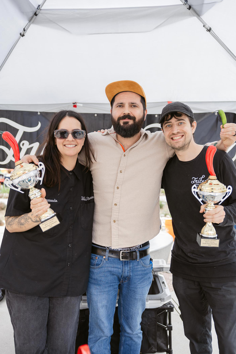 Three people standing under a canopy, with two holding trophies. The person on the left is a woman with dark hair, sunglasses, and tattoos, wearing a black shirt. The person in the middle is a man with a beard, wearing a tan cap, beige shirt, and jeans. The person on the right is a man with dark curly hair, wearing a black cap and black shirt. All are smiling.