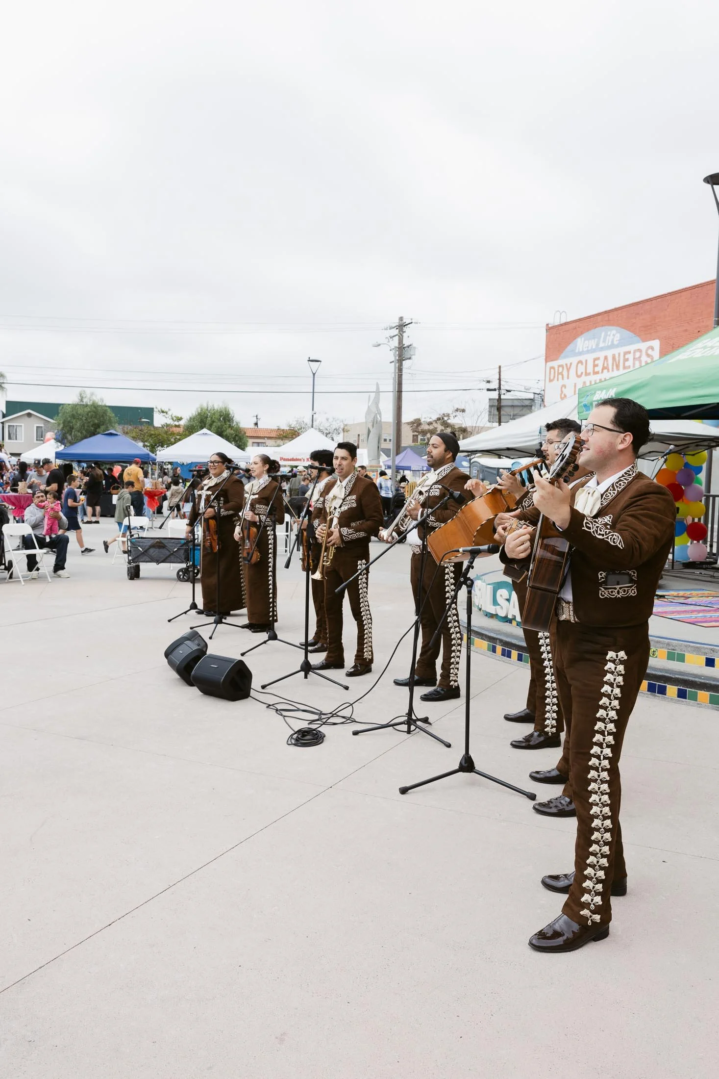 Musicians performing mariachi band at an outdoor event with audience, tents, and vendor stalls in the background.