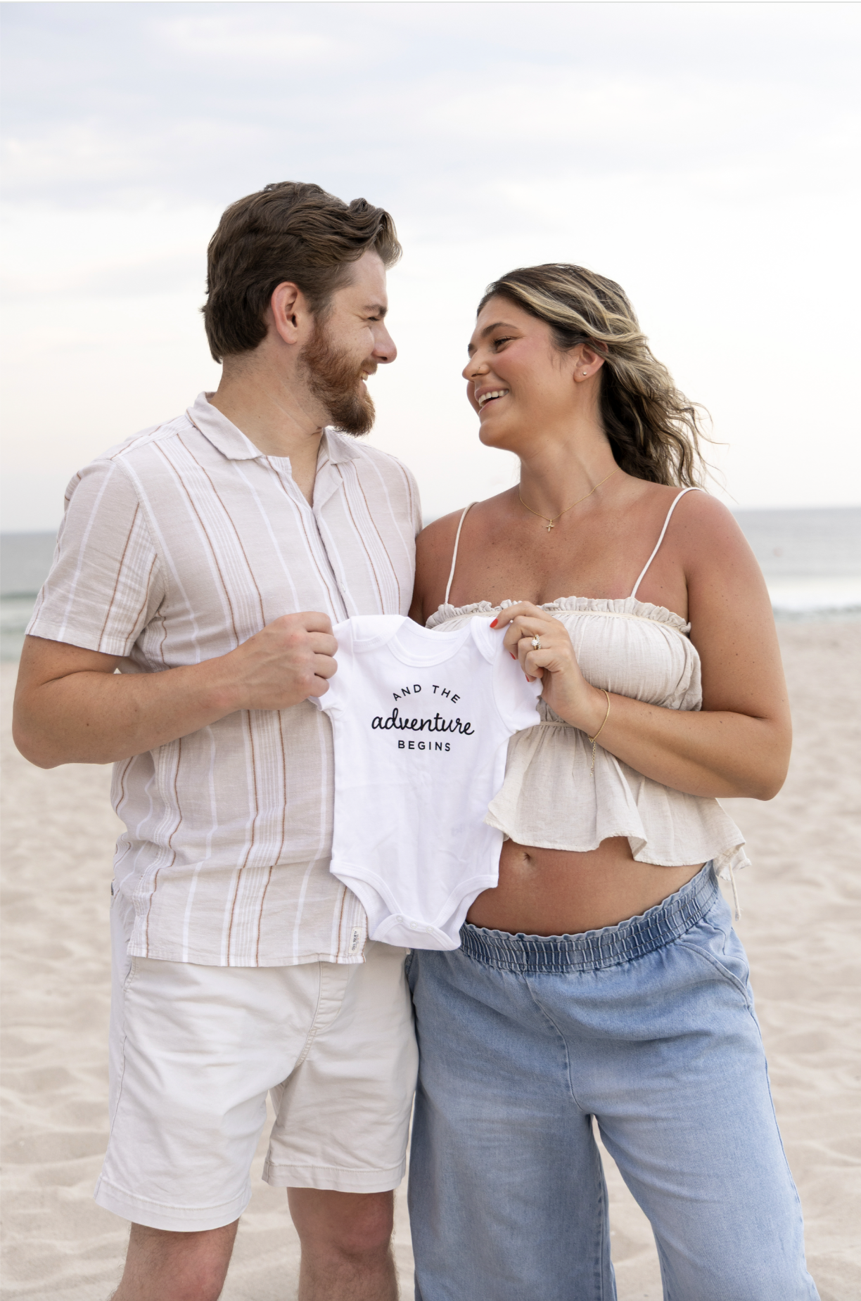 A smiling couple at the beach holding a white onesie that says 'and the adventure begins' between them, facing each other. jersey shore maternity photography