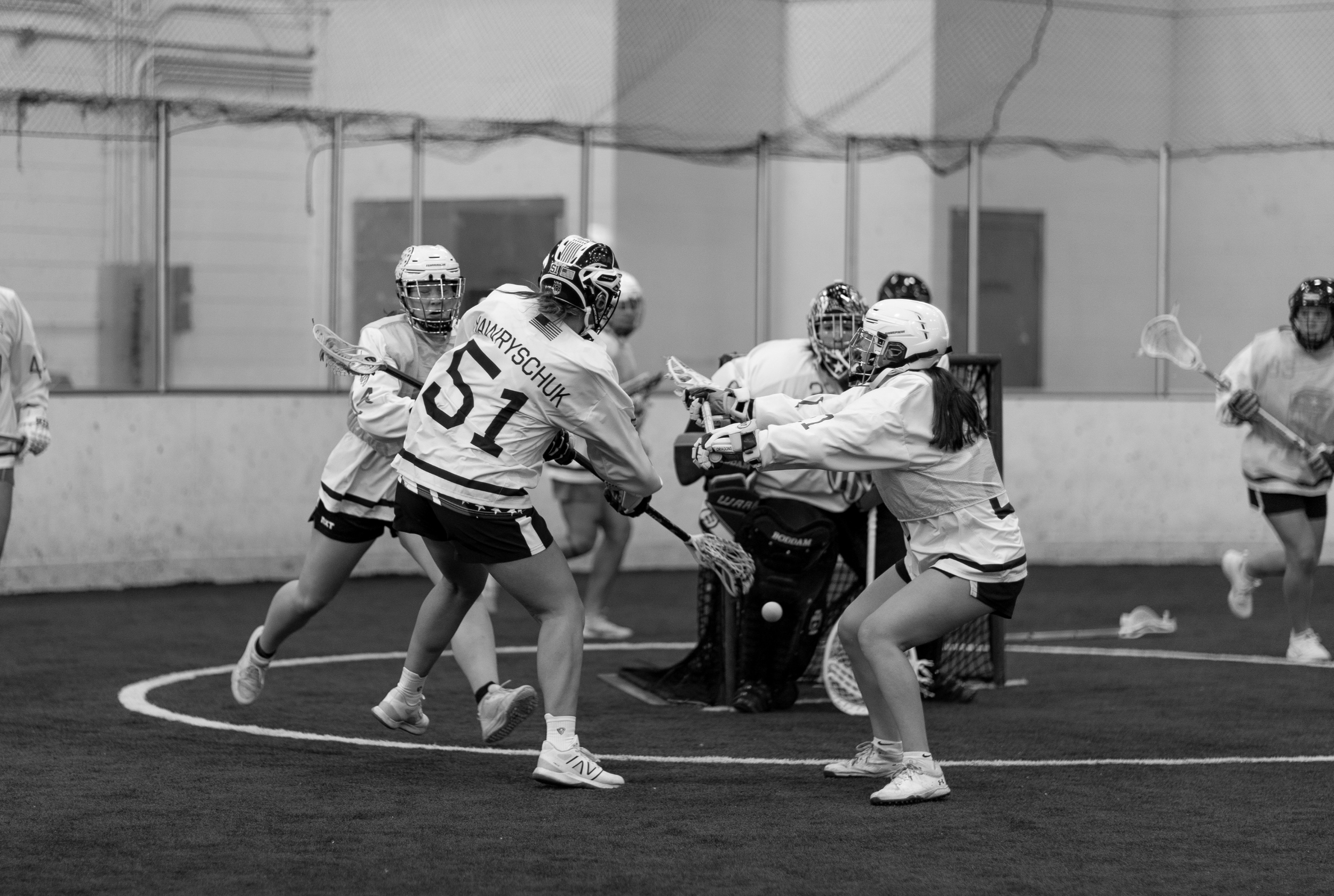 Women playing indoor lacrosse, with one trying to score and others defending, in an indoor sports facility.