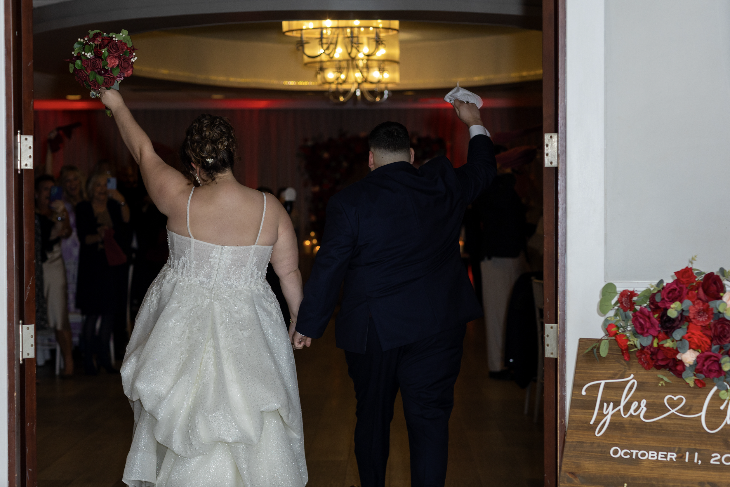 Bride and groom entering a wedding reception holding hands, with the bride waving a bouquet and the groom holding a napkin, surrounded by guests.