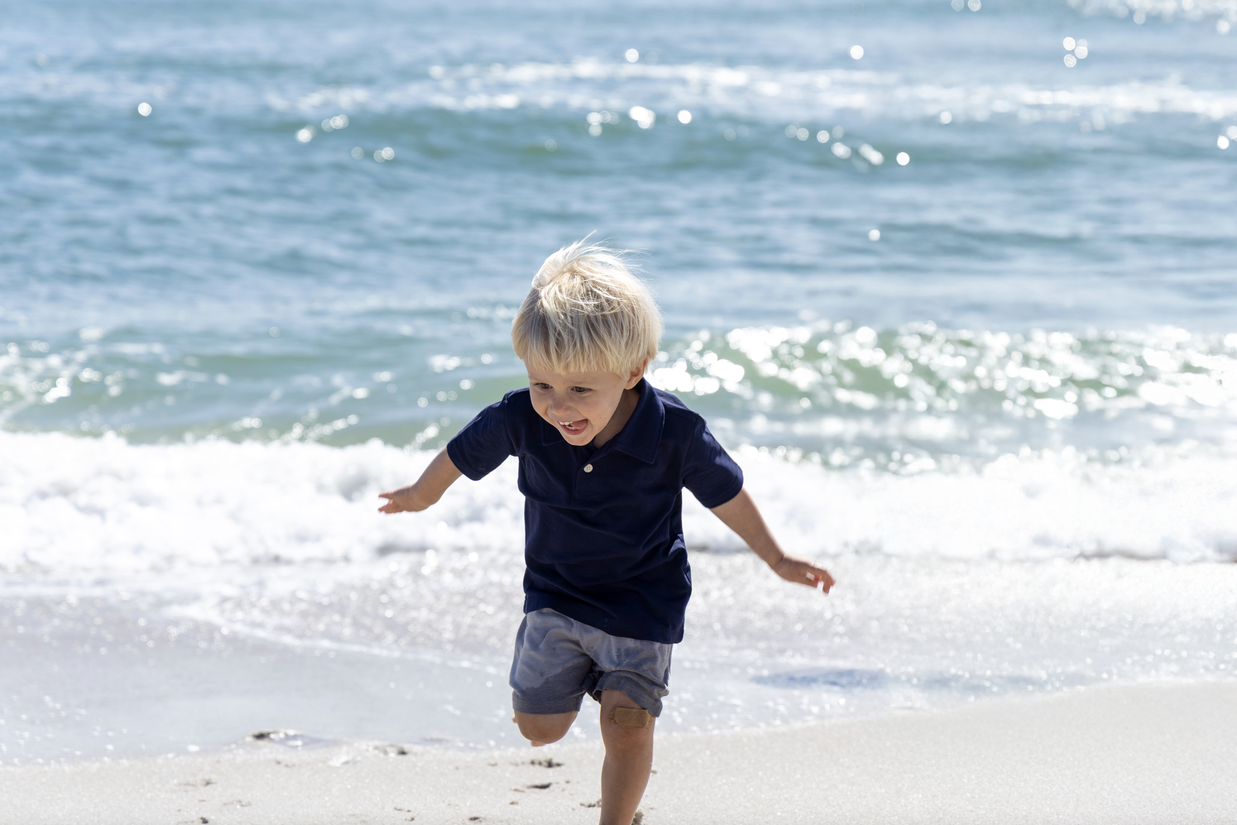 A young boy with blonde hair wearing a navy blue shirt and gray shorts running on the sandy beach near the ocean waves. Beach Family Photography New Jersey