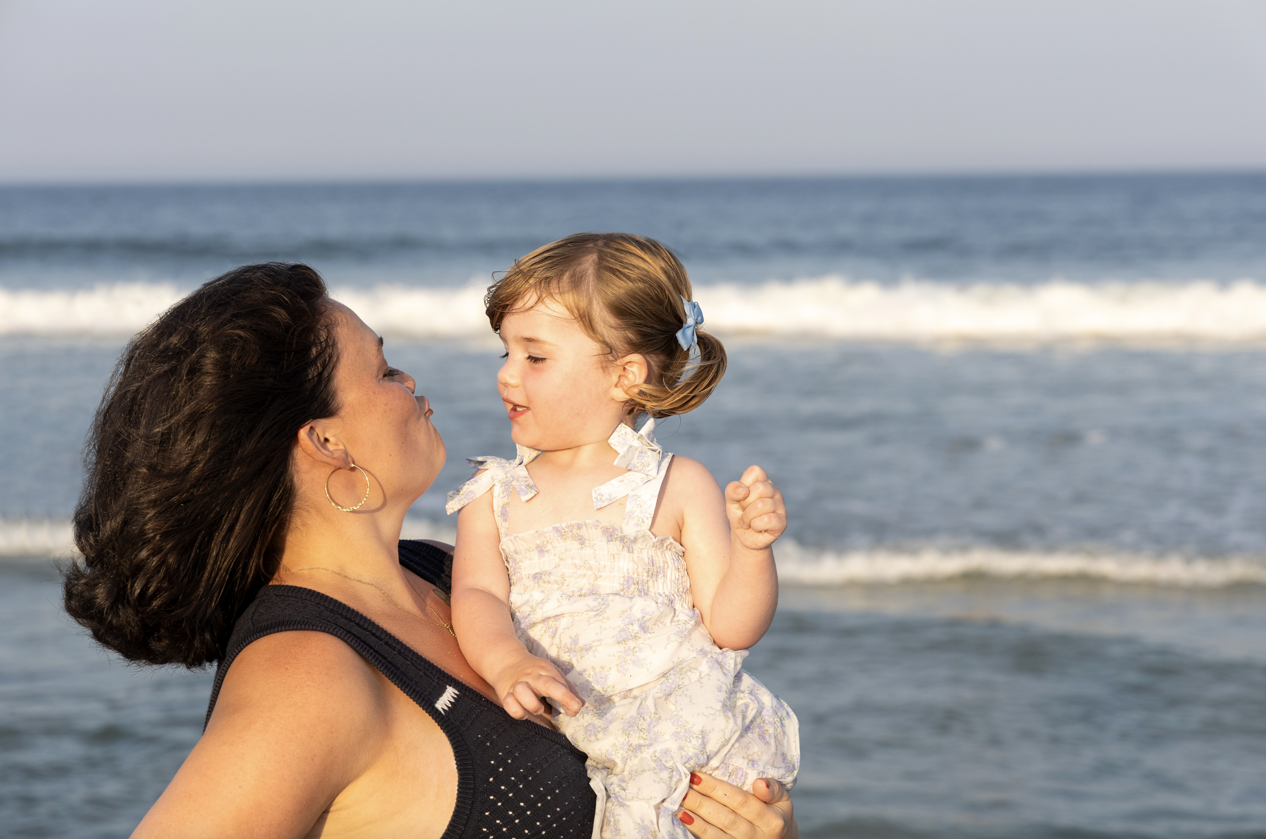 A woman holding a young girl in her arms on the beach with ocean waves in the background. Mom and Me Photography Lavallette, NJ