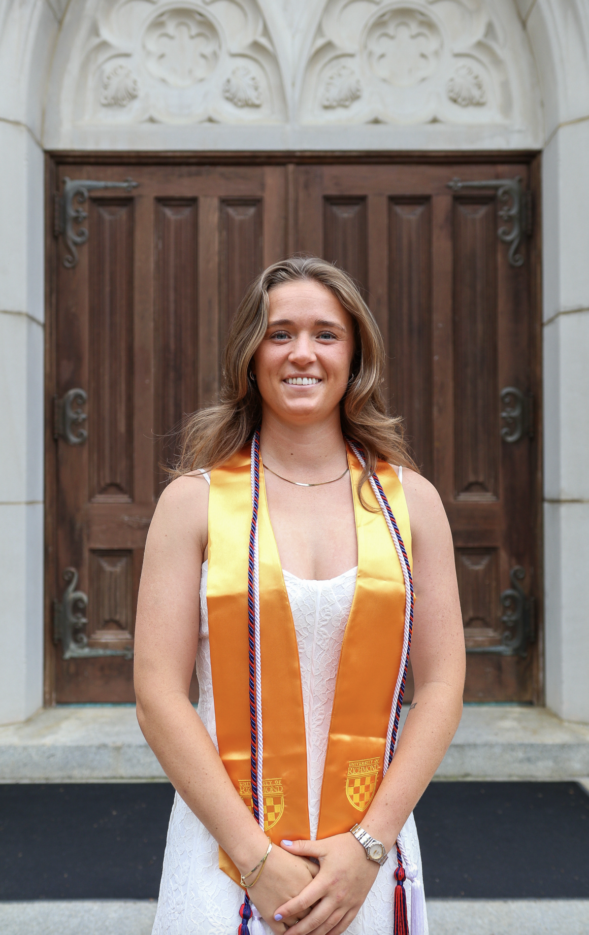 A young woman wearing a white dress and an orange graduation stole with cords stands in front of a large wooden door, smiling for a graduation photo.