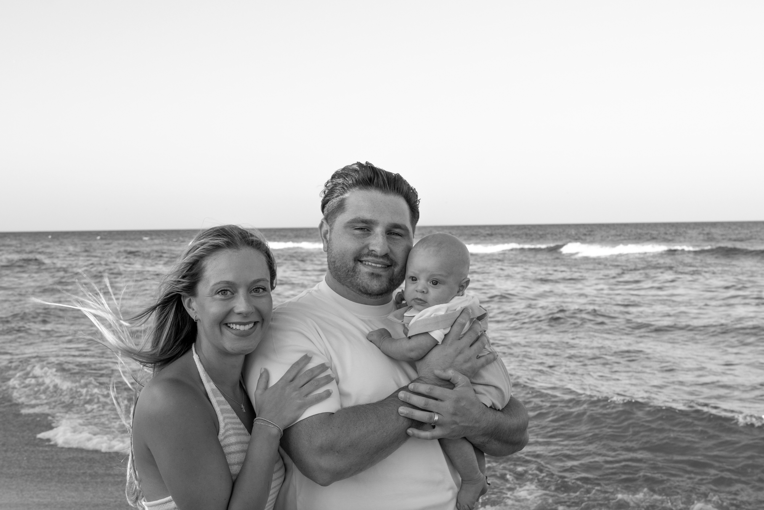 A family of three at the beach, a woman, a man, and a baby in the man's arms, smiling in front of the ocean.