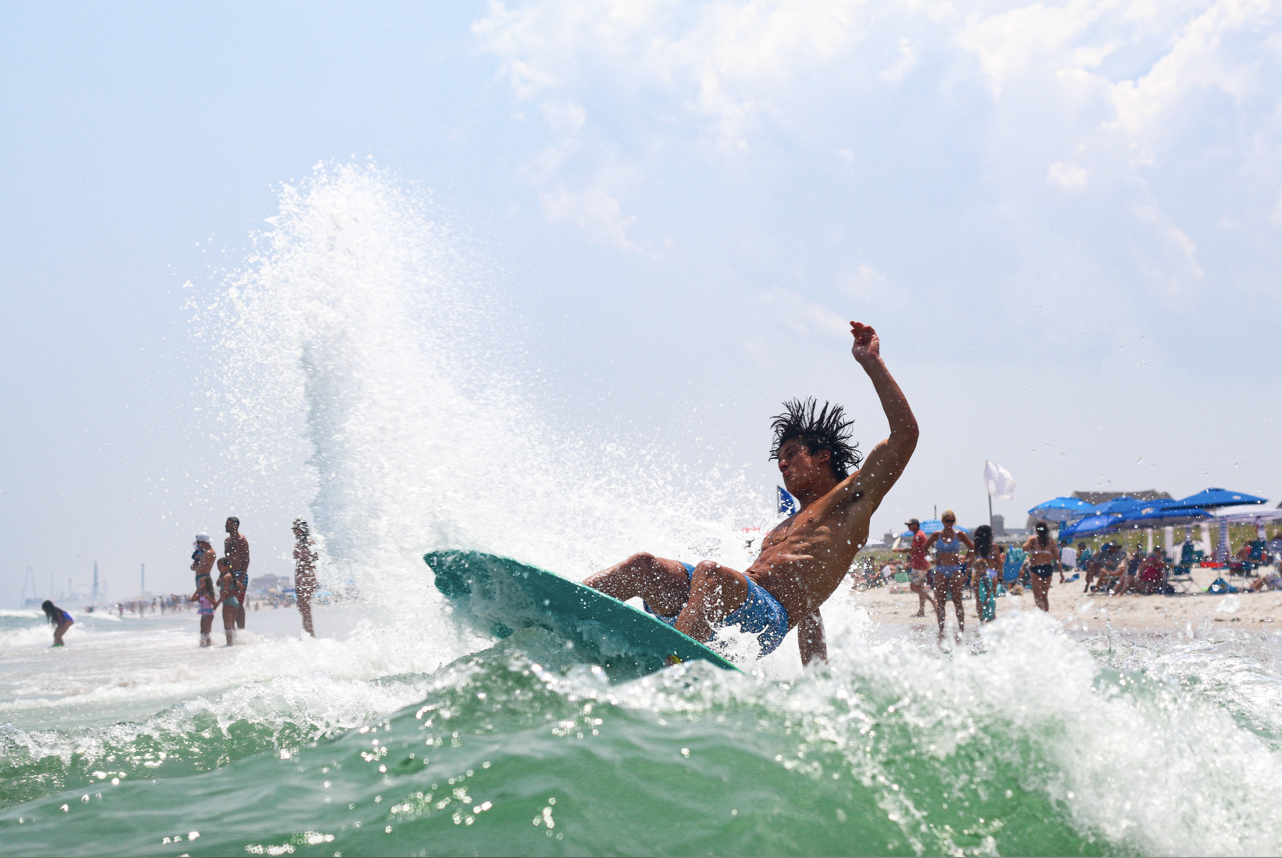 A man with long black hair surfing on a wave at the beach, with beach umbrellas and people in the background. Lavallette, NJ In water sports photography