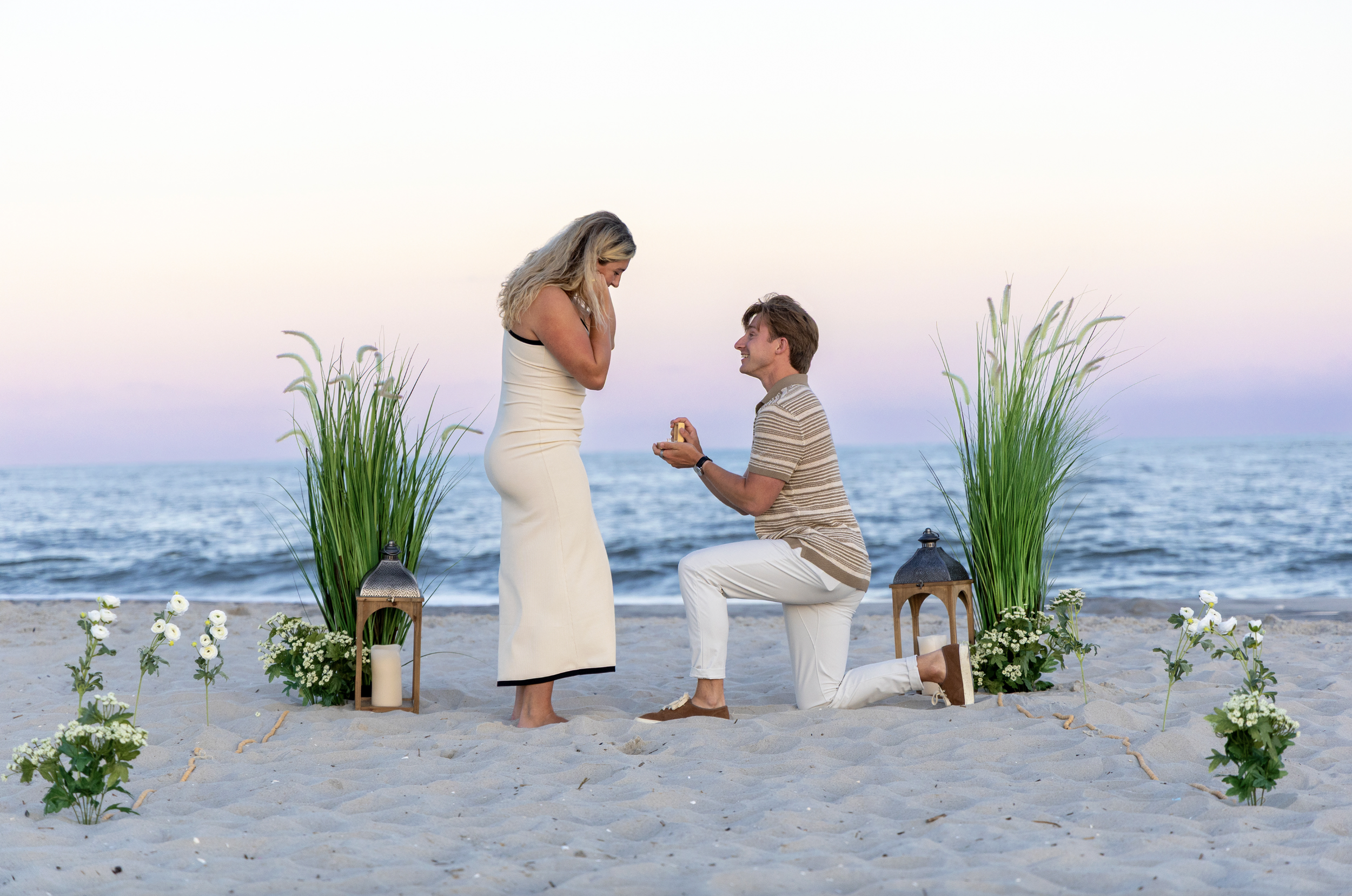 A man proposing to a woman on a beach during sunset, with the ocean in the background, surrounded by greenery and lanterns. In Cape May, New Jersey.