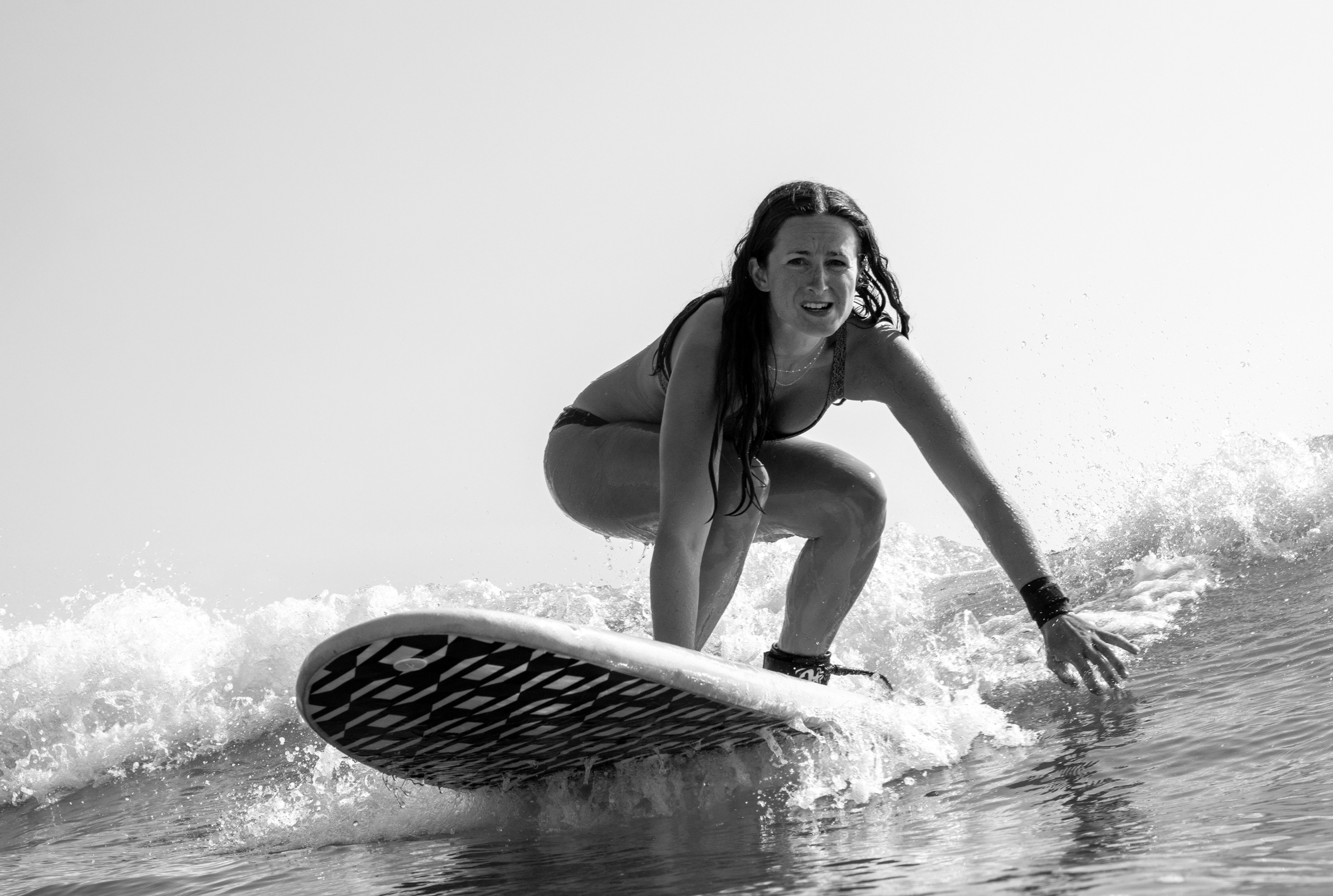 A woman with long hair crouches on a surfboard riding a wave at the beach in black and white.