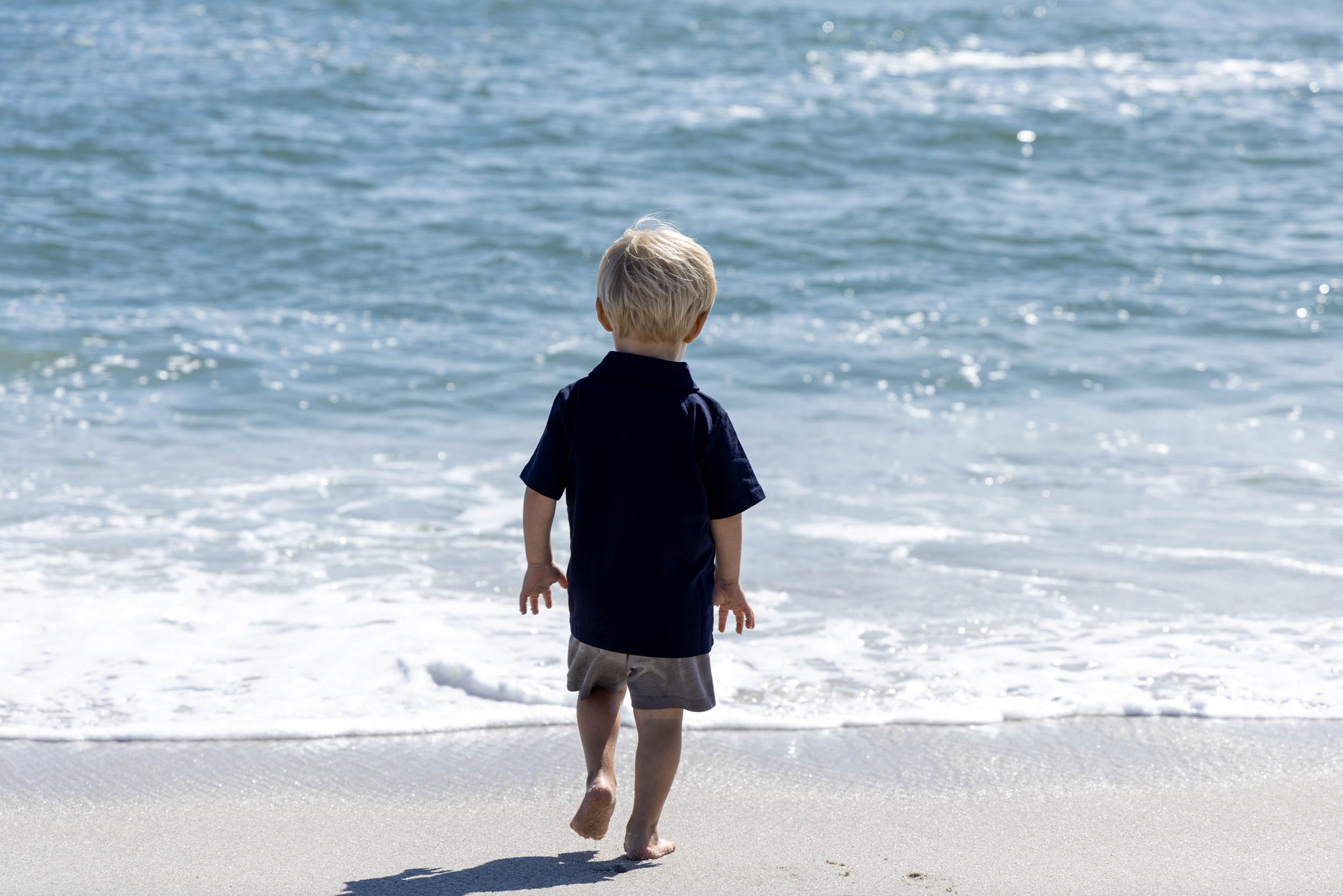 A young boy with blonde hair, wearing a navy blue polo shirt and gray shorts, walking barefoot on the sandy beach toward the ocean.