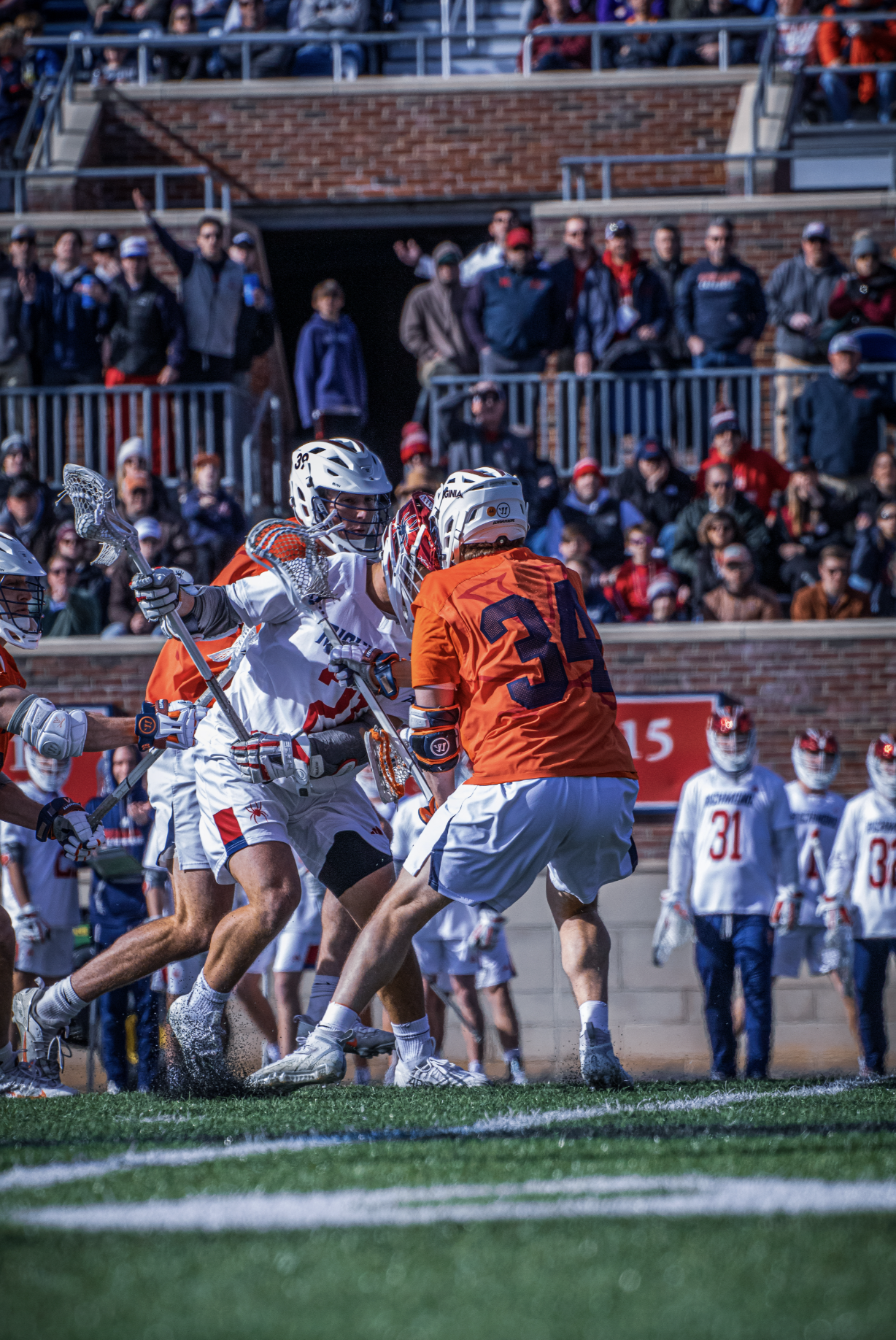 Lacrosse players in white and orange jerseys collide during a game on a green field, with spectators watching from the stands in the background.